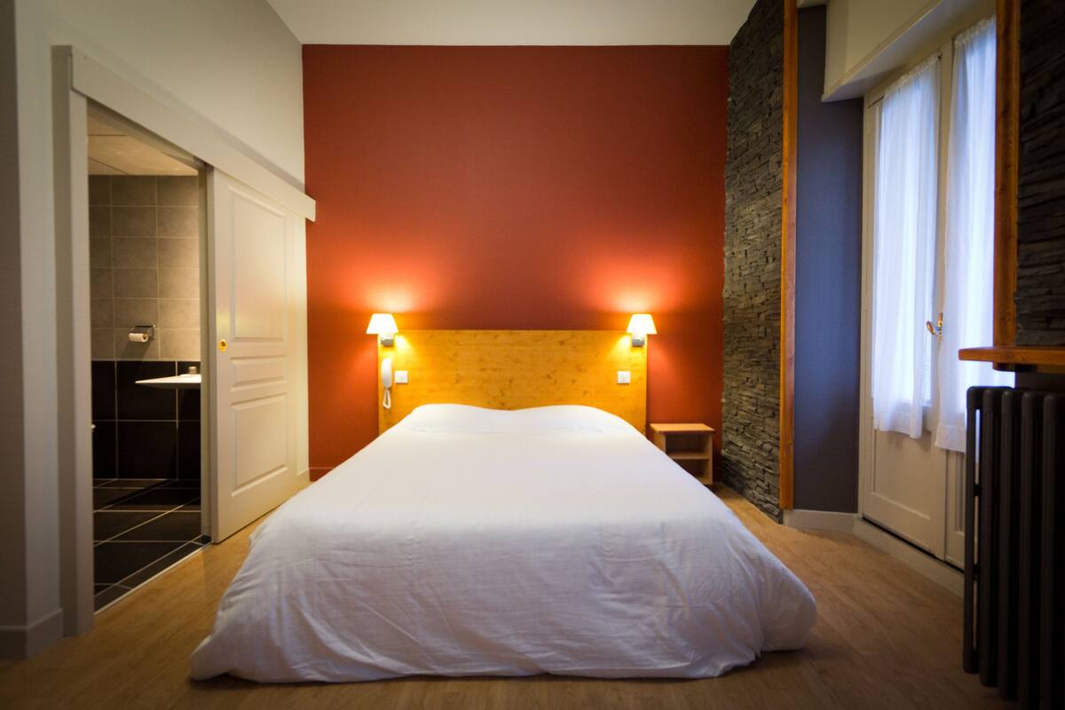 Quarto de hotel com cama grande, duas luminárias na parede, cabeceira de madeira, parede de destaque em pedra, radiador e porta para um banheiro.