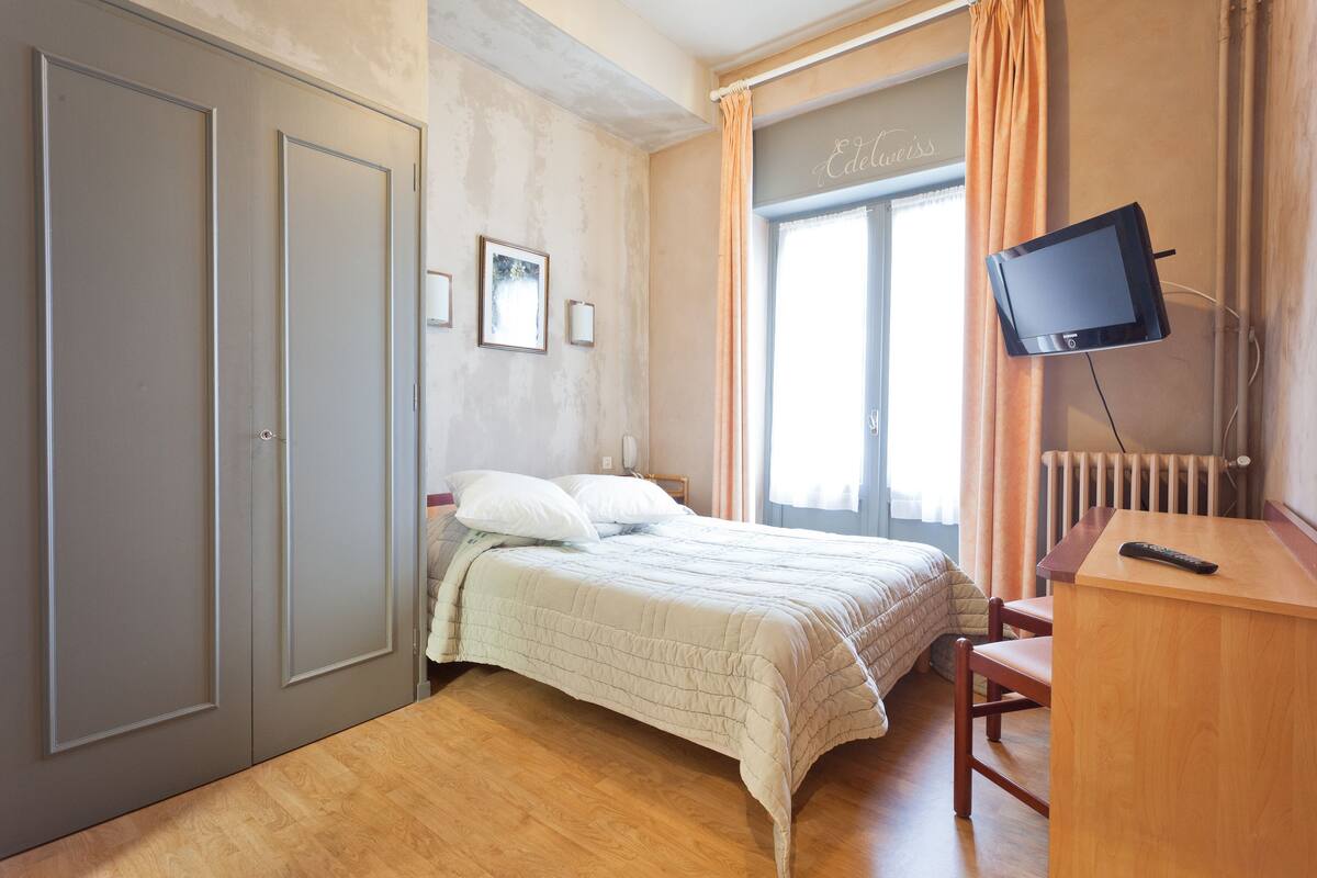 Quarto com cama, escrivaninha, televisão e janela com cortinas.