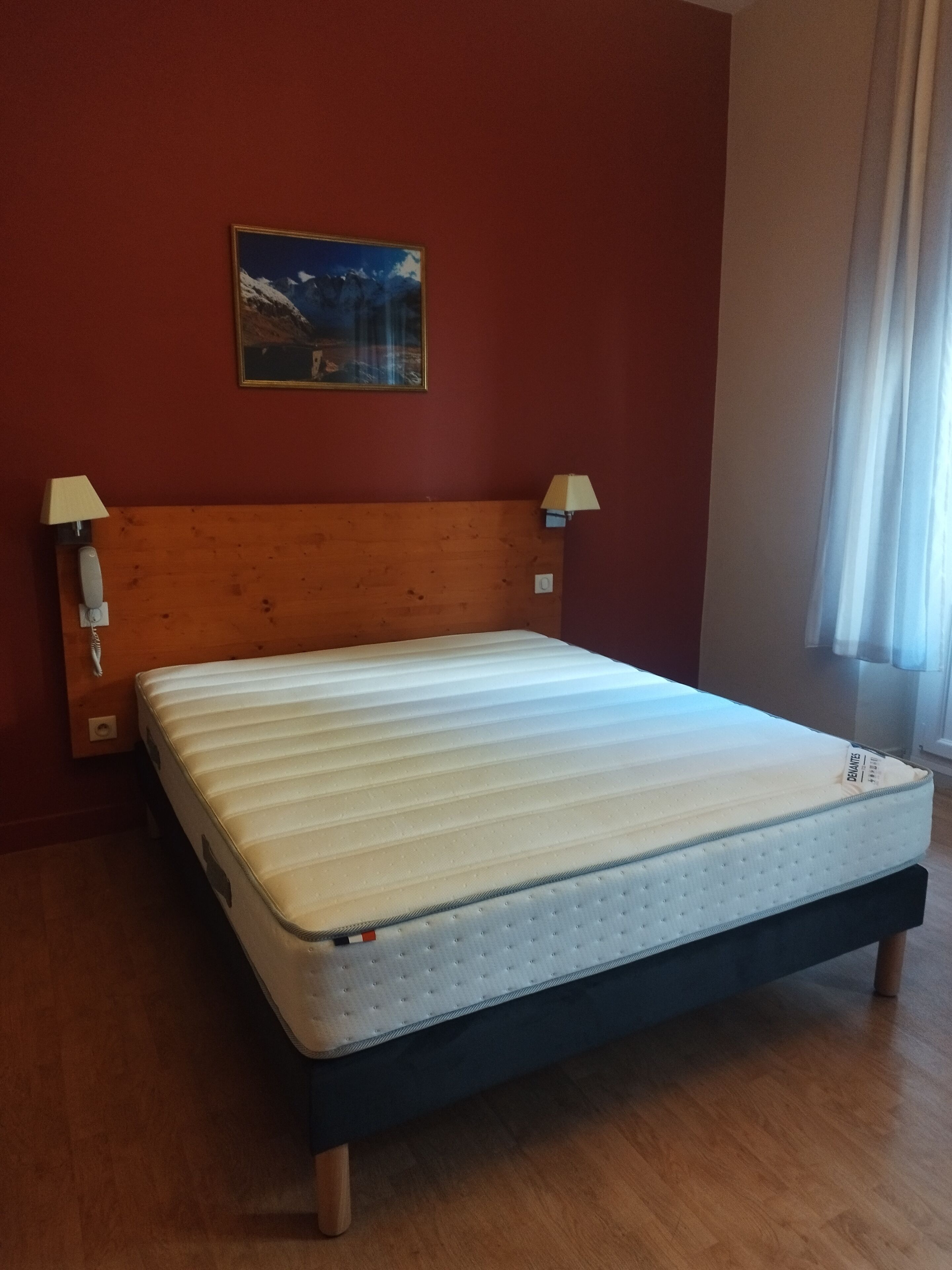 Une chambre avec un lit doté d’une tête de lit en bois, deux lampes de chevet, un tableau au mur et une fenêtre avec des rideaux.