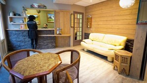 Zona de recepción con recepcionista, mostrador, sofá amarillo y una mesa de comedor con sillas.