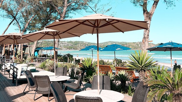 Un patio al aire libre con sillas y mesas de mimbre, sombrillas azules y vista a la playa y el océano.