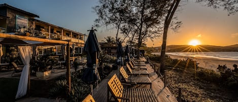 A wooden deck with lounge chairs and umbrellas, overlooking a beach at sunset.
