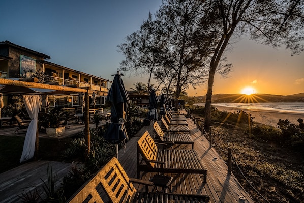 A wooden deck with lounge chairs and umbrellas, overlooking a beach at sunset.