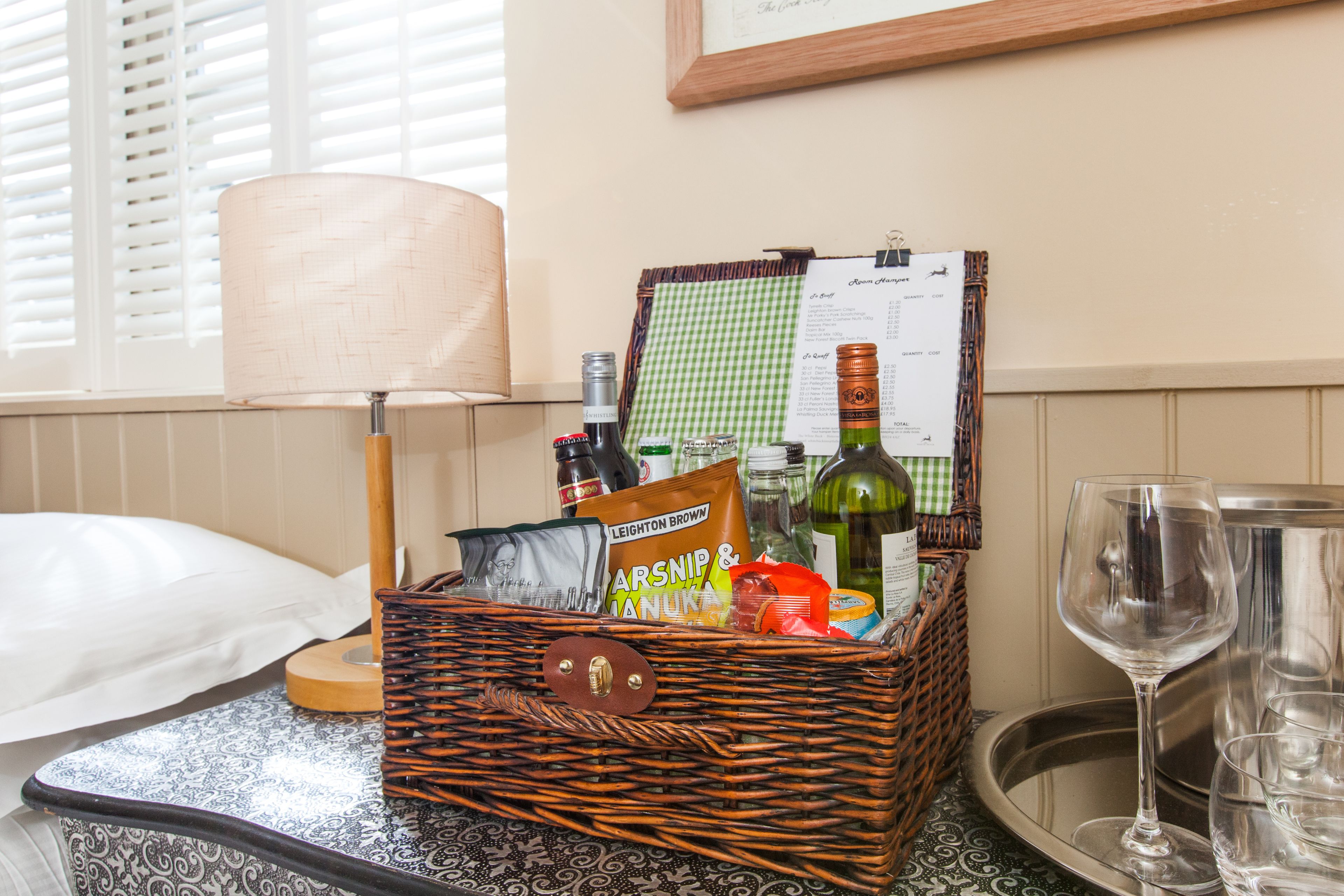 Una cesta de mimbre llena de aperitivos y bebidas sobre una mesa junto a una lámpara y copas de vino.
