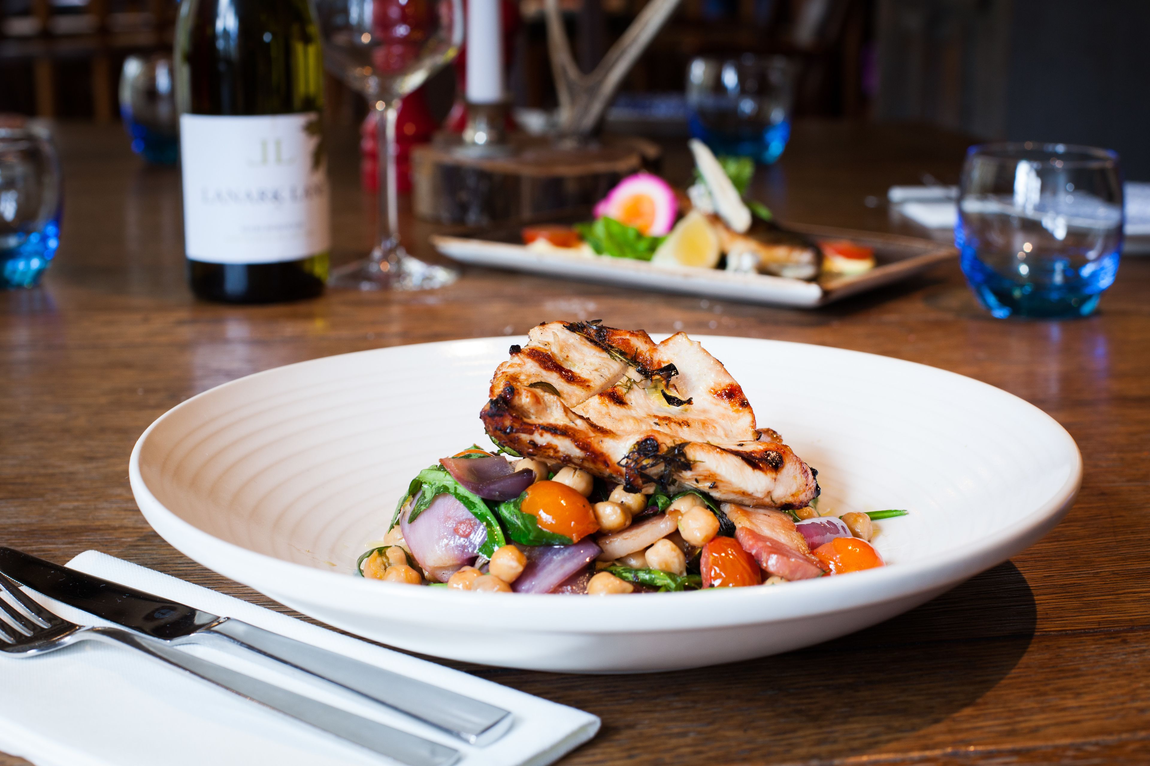 A plate of grilled chicken with vegetables and chickpeas on a white plate, set on a wooden table with a wine bottle and glasses in the background.