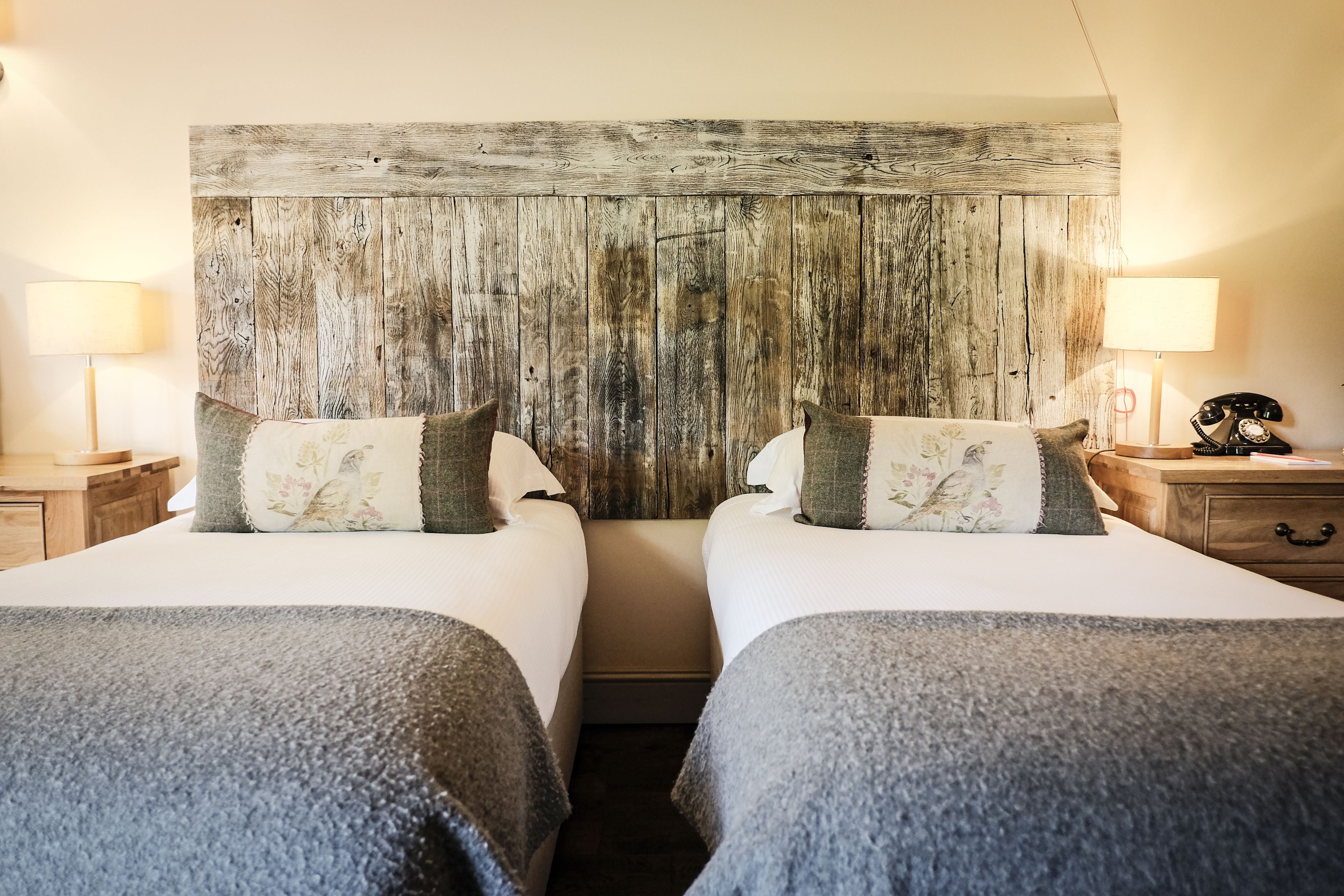 A double room with two beds, each with a grey blanket and floral pillows. A rustic wooden headboard separates the beds. There are bedside tables with lamps and a telephone on the right side.