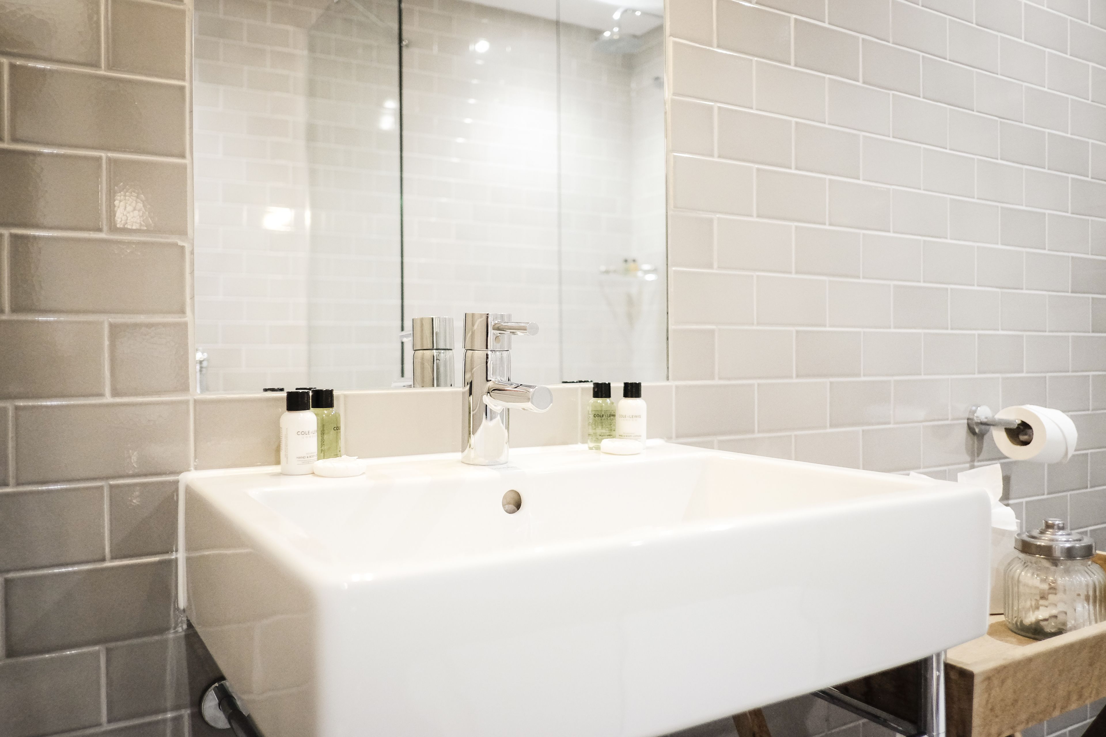A bathroom with a white sink, two faucets, and a mirror. There are toiletries on the sink and a glass jar on the countertop.
