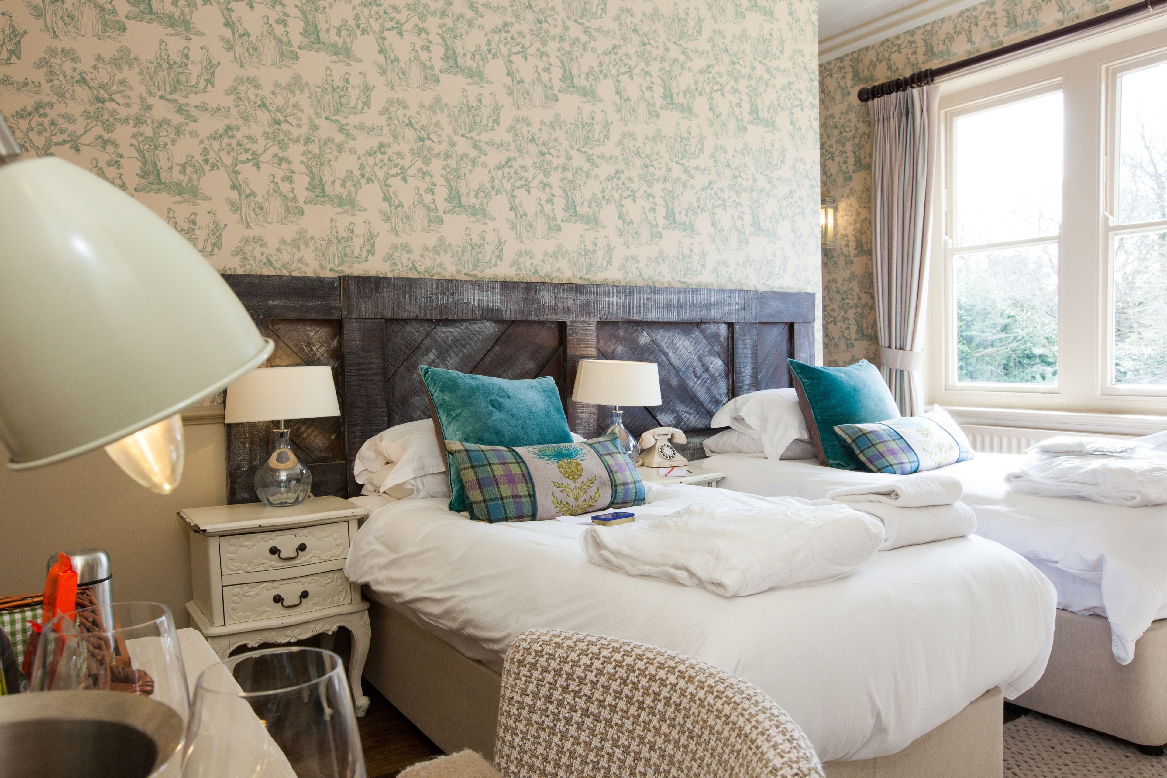 A hotel room with two beds, a nightstand with a lamp, a telephone, and a window with curtains.