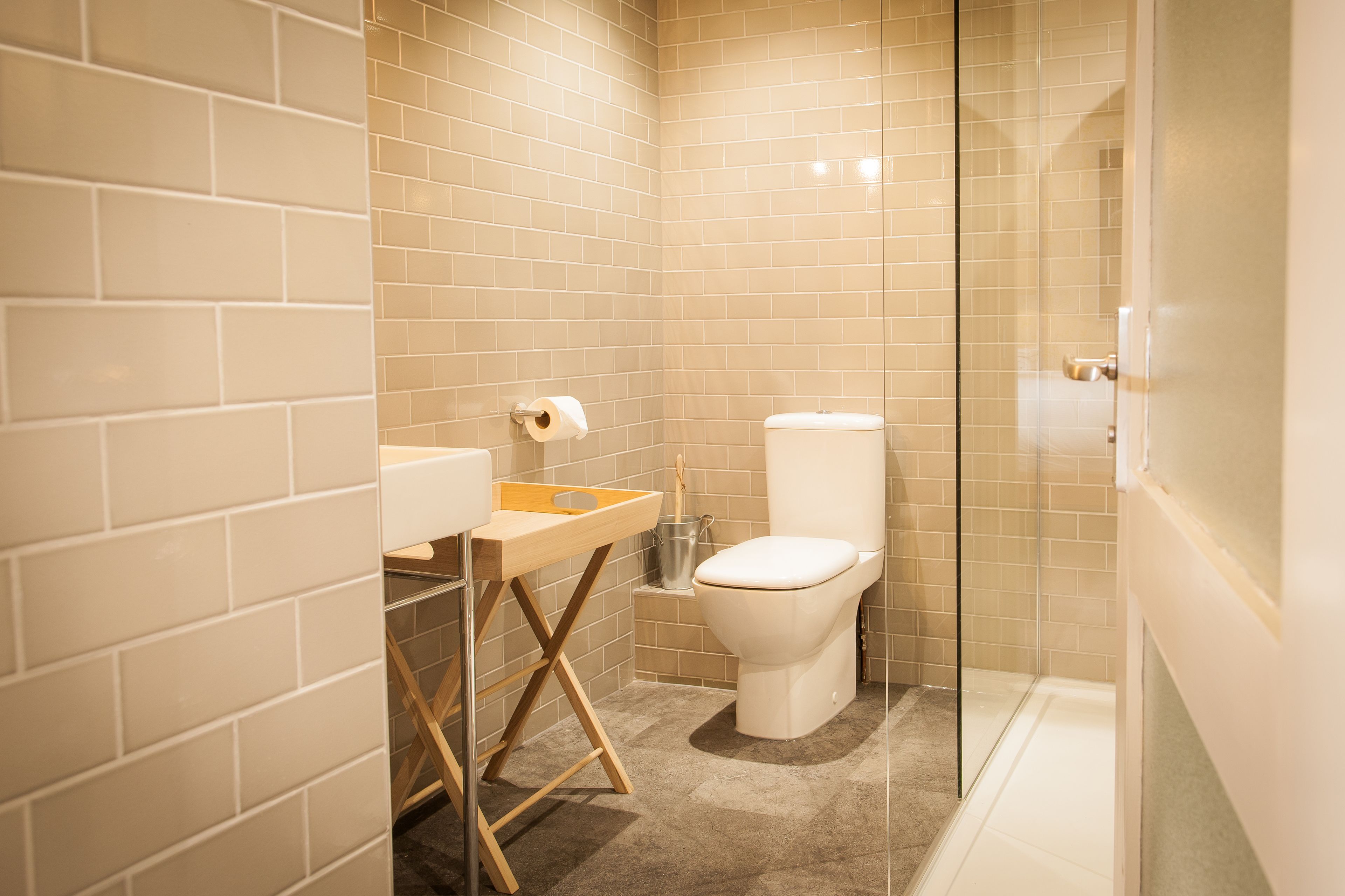 Een moderne badkamer met beige tegels, een wit toilet, een houten plank en een douchecabine met glazen wanden.