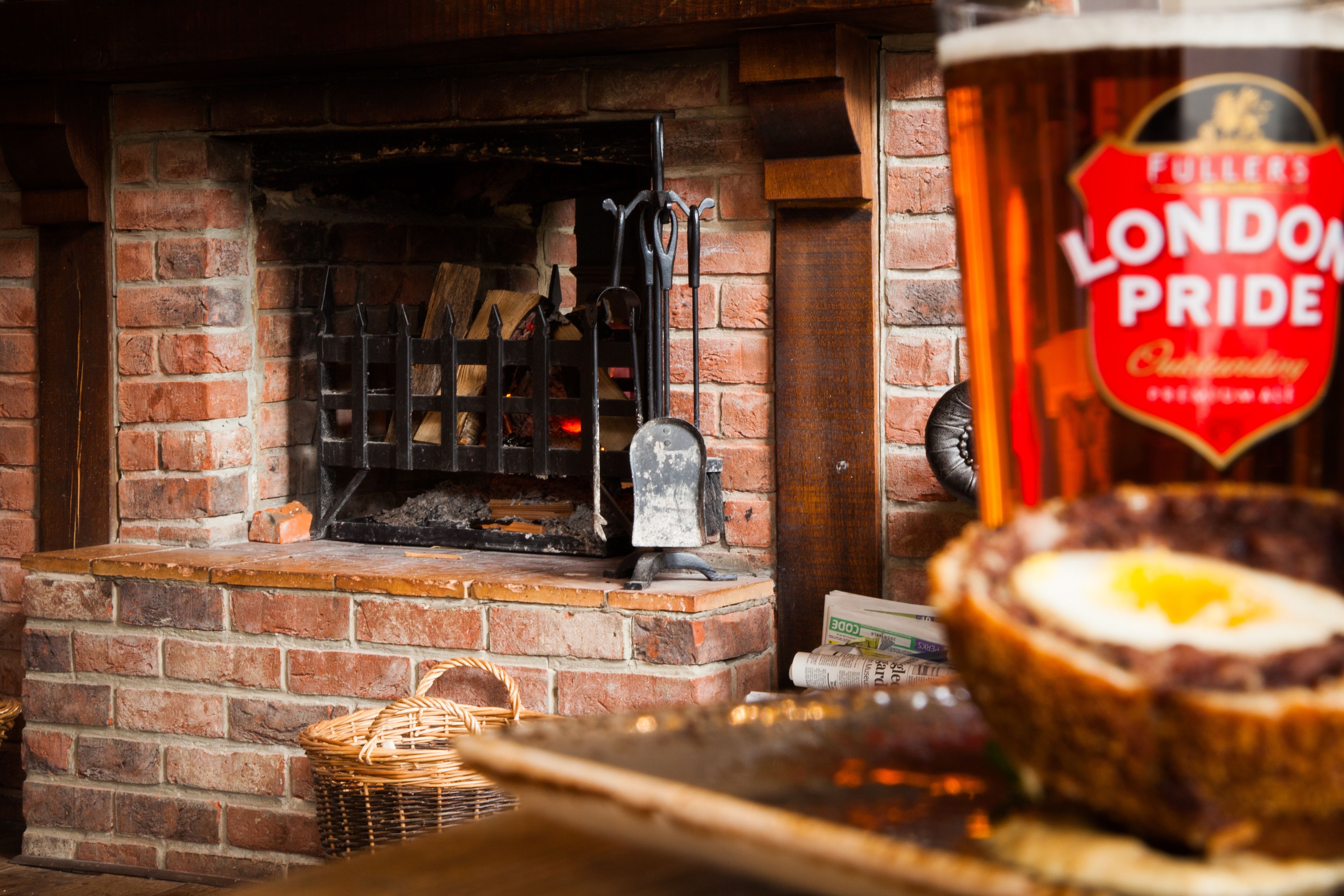 A brick fireplace with a log fire, a beer tap labeled "Fuller's London Pride," and a basket on the floor.