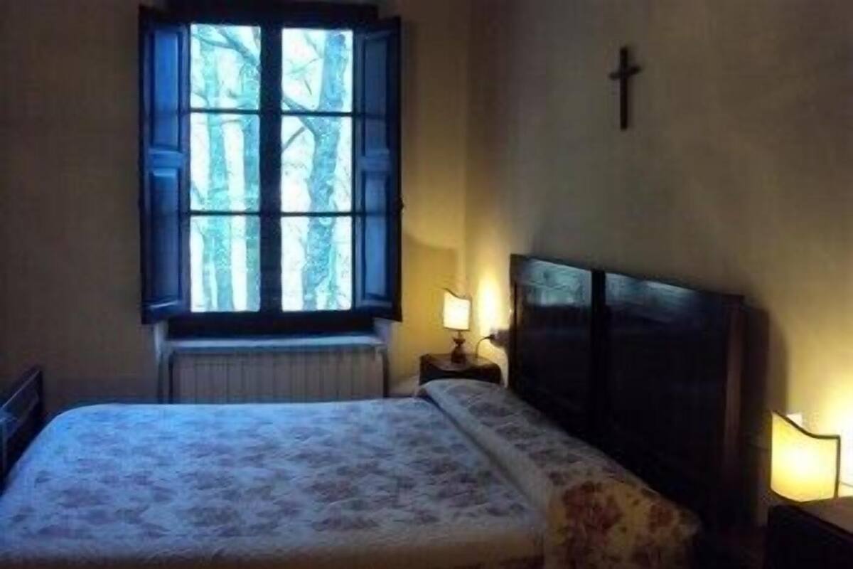 A bedroom with a bed, a nightstand, a window with a view of trees, and a cross on the wall.
