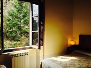 A bedroom with a large window, a radiator, a bed with a floral bedspread, and a bedside lamp. - Locanda dei Baroni (Poppi)