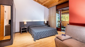 A hotel room with a wooden ceiling, a bed with a floral bedspread, a nightstand with a lamp, a seating area with a cushion, and a sliding glass door to the outside.