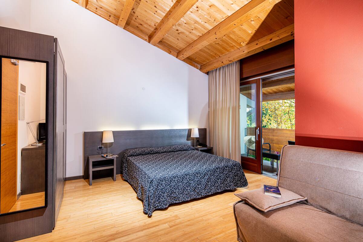 Ein Hotelzimmer mit Holzboden, einem Bett mit floralem Überwurf, einem Nachttisch mit Lampe, einer Sitzecke mit Kissen und einer Schiebetür zum Außenbereich.