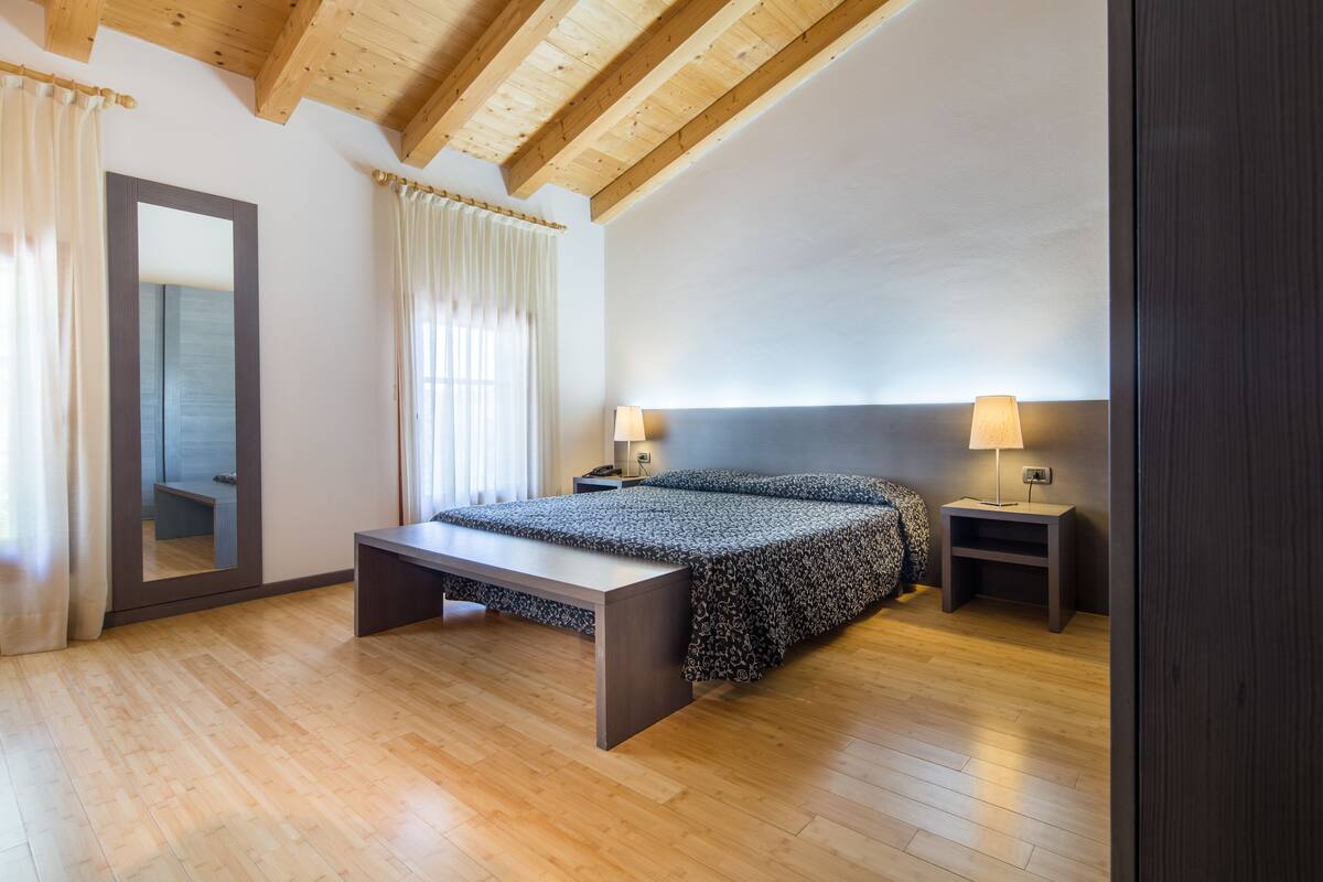 Ein Schlafzimmer mit Holzboden, einem Bett mit gemustertem Überwurf, einem Nachttisch mit Lampe, einem großen Spiegel und einem Fenster mit Vorhängen.