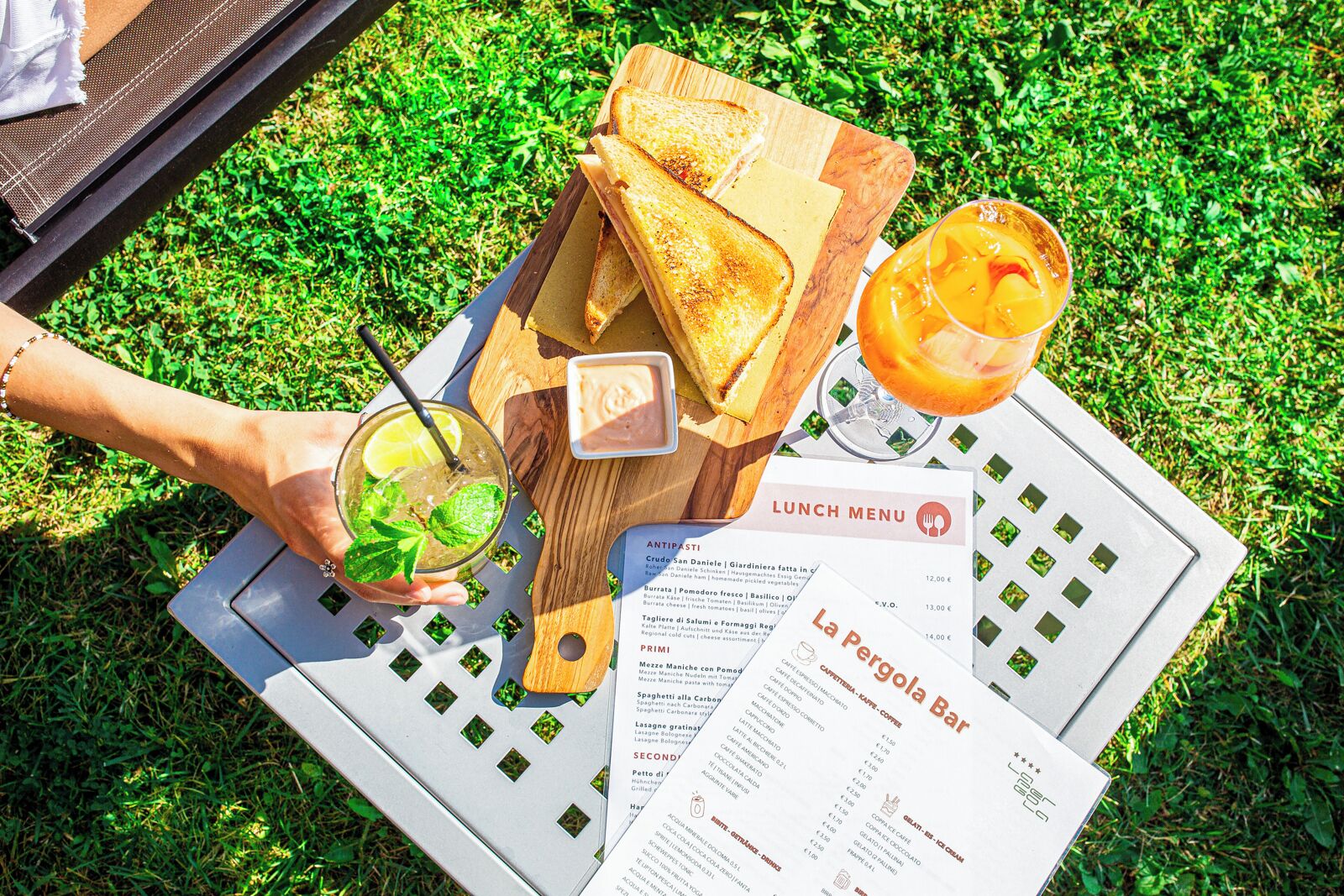 A lunch menu on a table with a wooden board holding toasted bread, a drink with a lemon slice, and a menu with food items listed.