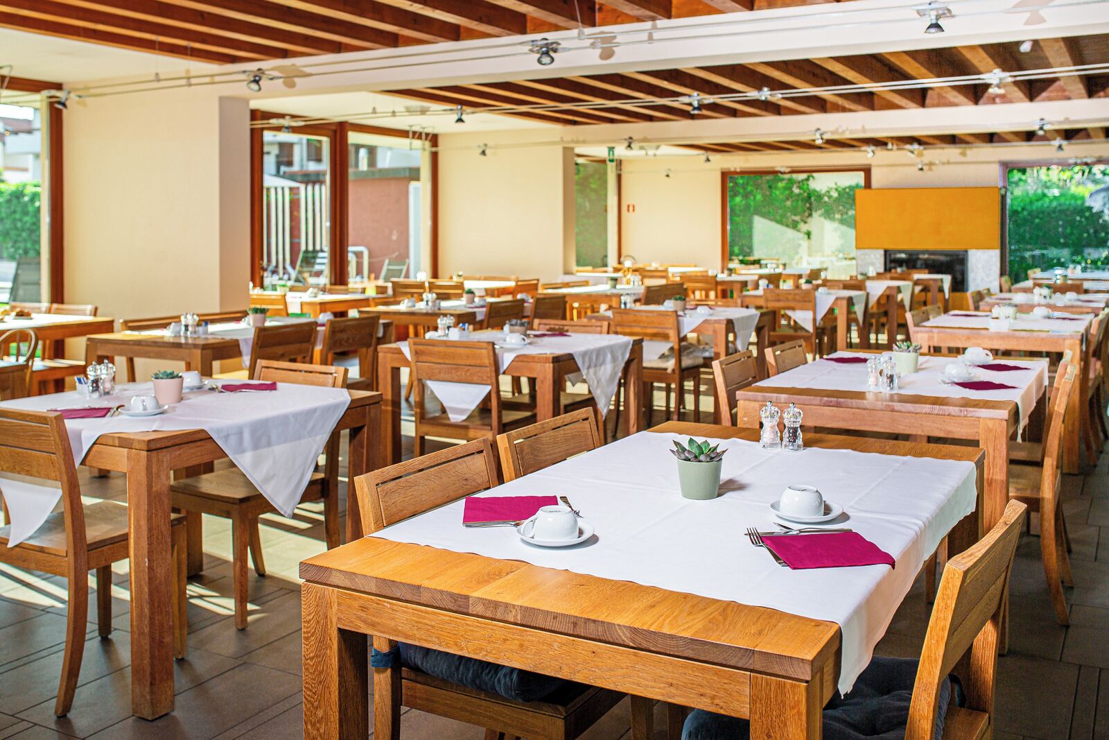 Un amplio comedor con varias mesas y sillas de madera, manteles blancos y pequeñas plantas en macetas.