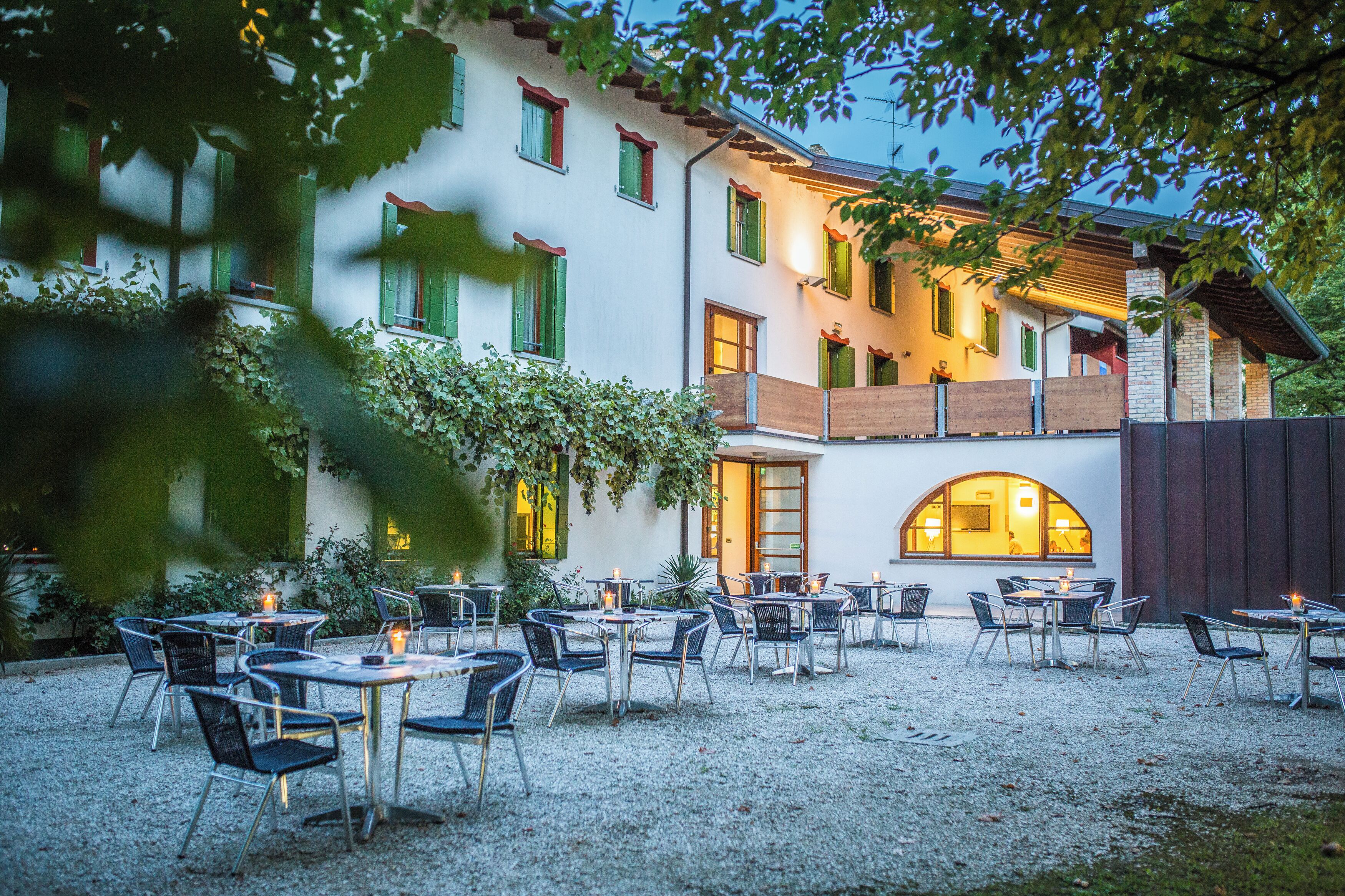 An outdoor cafe with tables and chairs, surrounded by a building with multiple windows and a gravel ground.