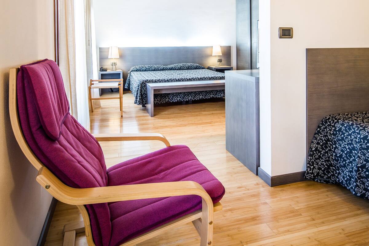 Ein modernes Hotelzimmer mit Holzboden, einem Bett mit gemustertem Überwurf, einem Nachttisch mit Lampe und einem Sessel mit violettem Kissen.