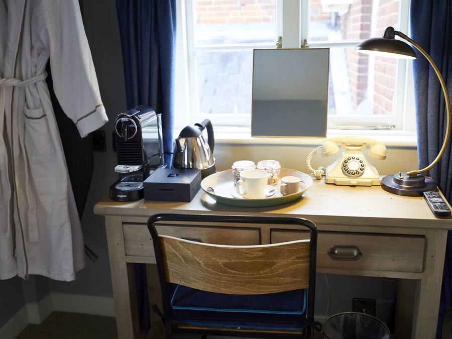 Una camera d'albergo con una scrivania in legno, una sedia, un accappatoio appeso, un bollitore, tazze, un telefono e una lampada.