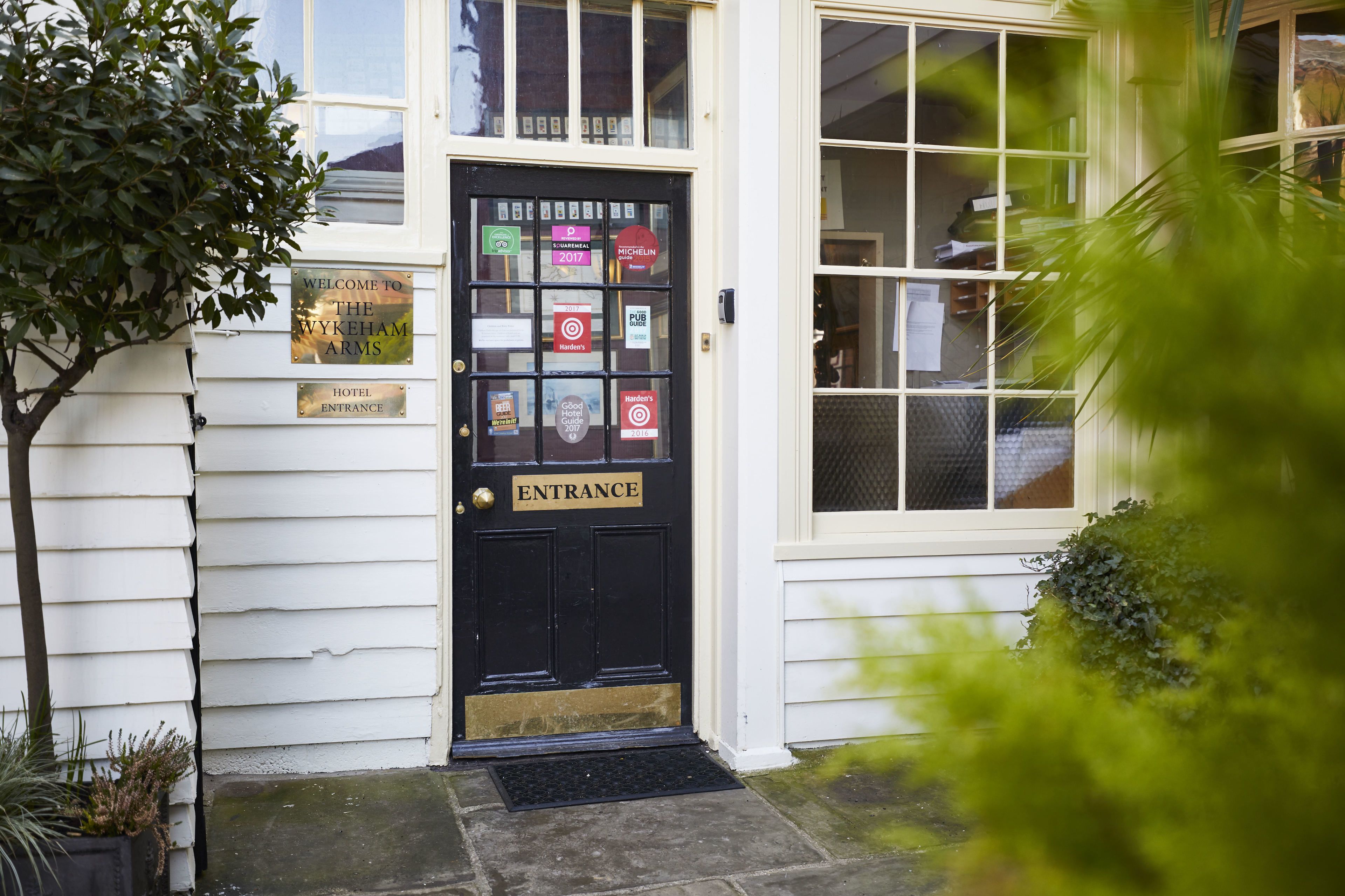 A black entrance door with a 'Welcome to the Wykeham Arms' sign, a 'Hotel Entrance' sign, and various awards and certifications.