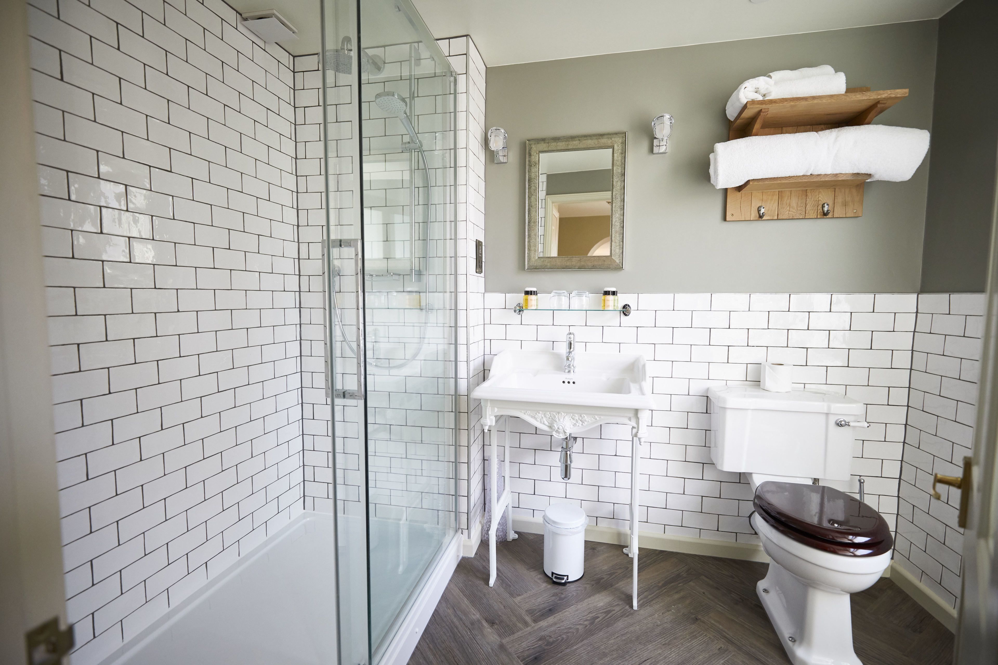 Een badkamer met witte metrotegels, een glazen douchecabine, een wit wastafelmeubel, een toilet en een houten plank met handdoeken.