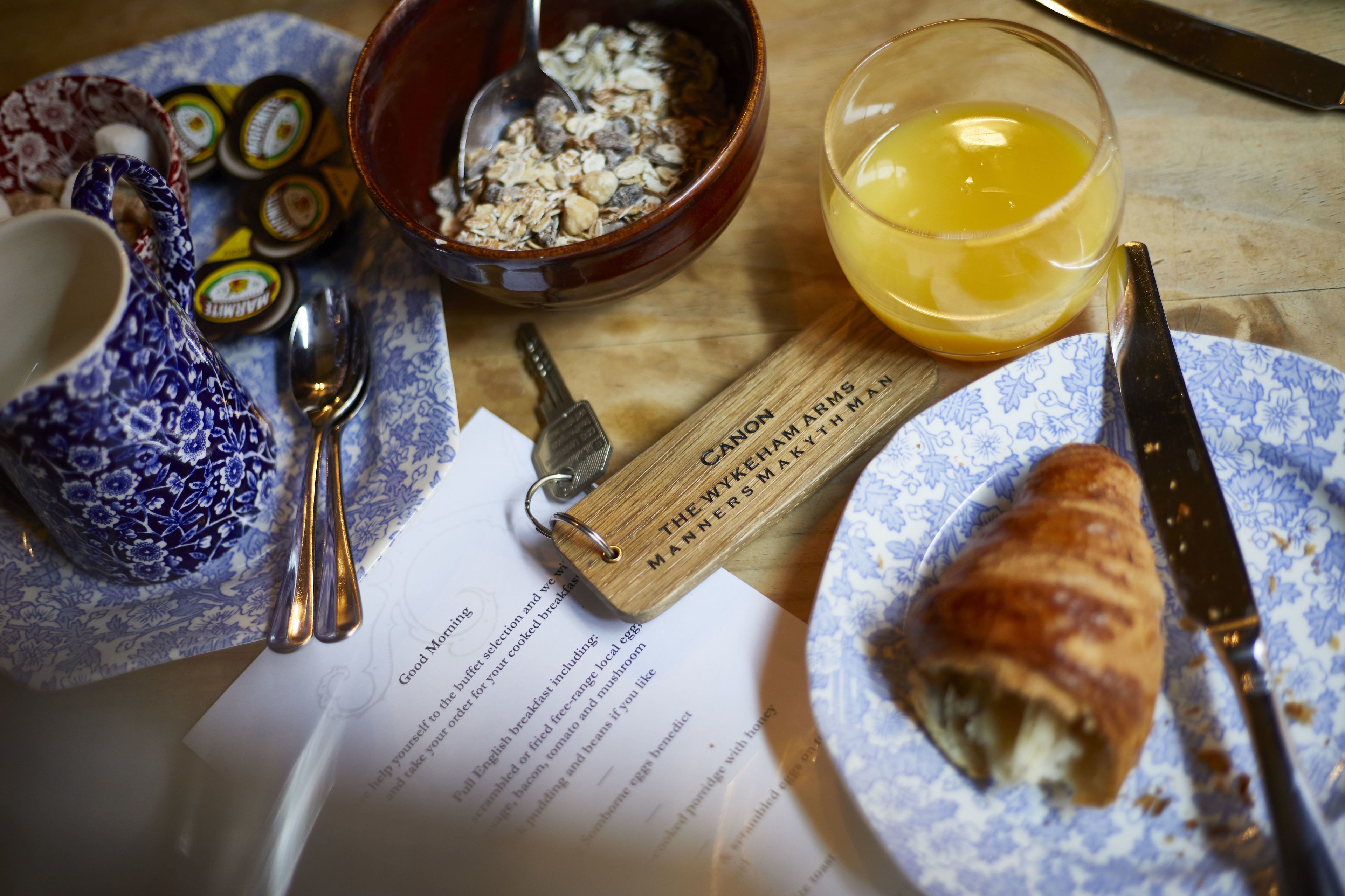 Un desayuno con un croissant, un vaso de zumo de naranja, un bol de cereales, una cuchara, un cuchillo, una tetera y un llavero.
