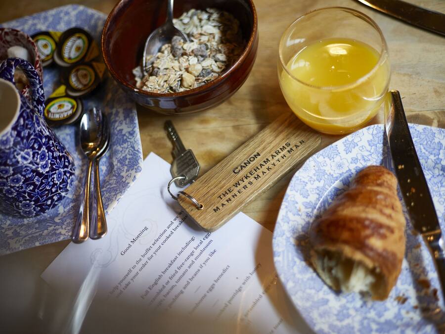 Un desayuno con un croissant, un vaso de zumo de naranja, un bol de cereales, una cuchara, un cuchillo, una tetera y un llavero.