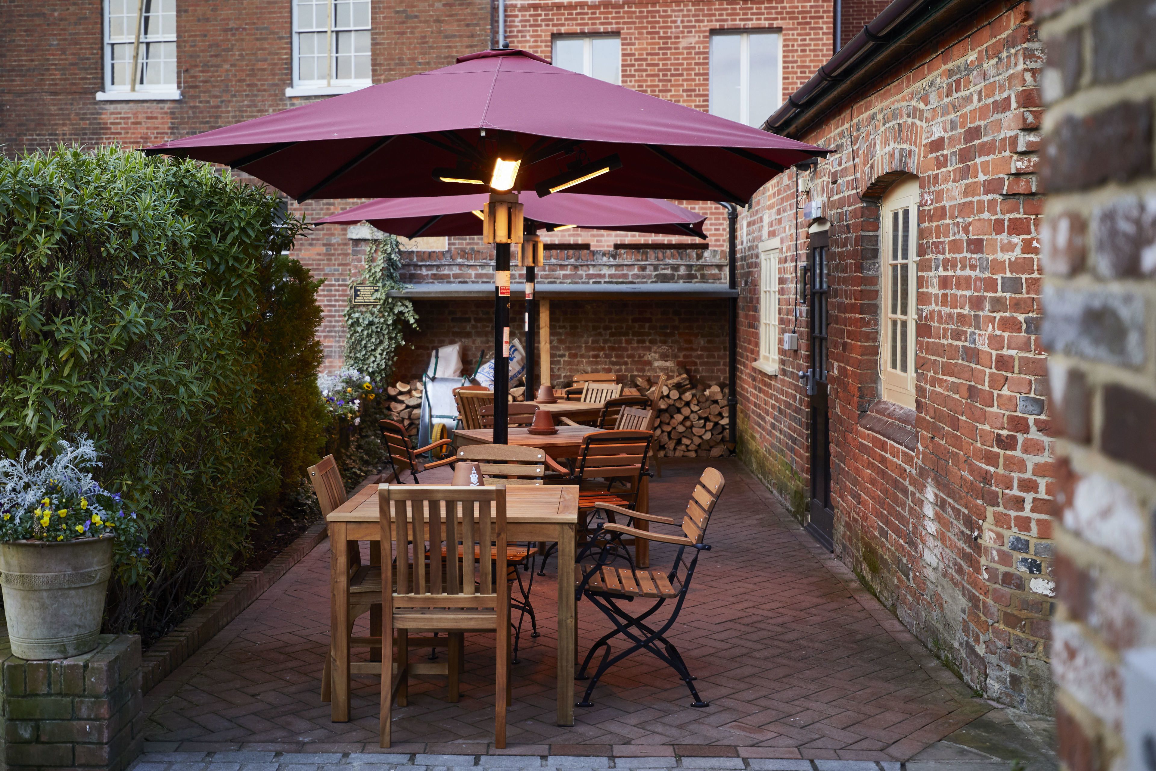 An outdoor patio with wooden tables and chairs, a large umbrella, and a brick building in the background.