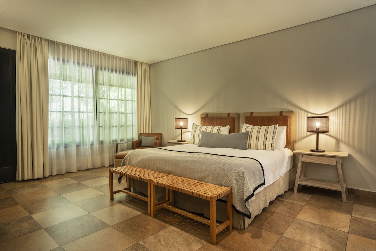 Habitació d'hotel amb un llit gran, dues taules de nit amb làmpades, una panxa i una finestra gran amb cortines.