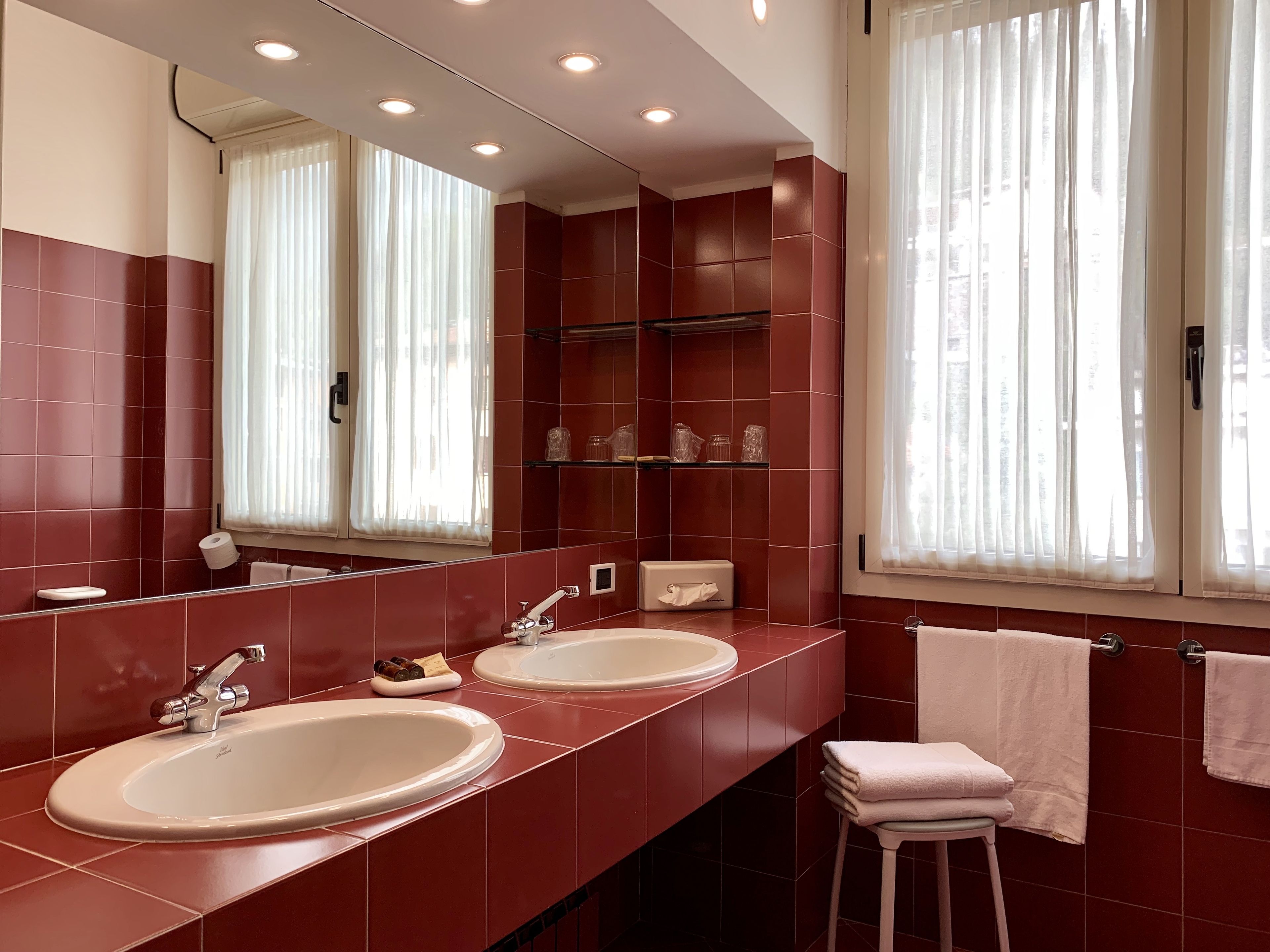 Un baño moderno con dos lavamanos, un espejo grande y una ventana con cortinas translúcidas.