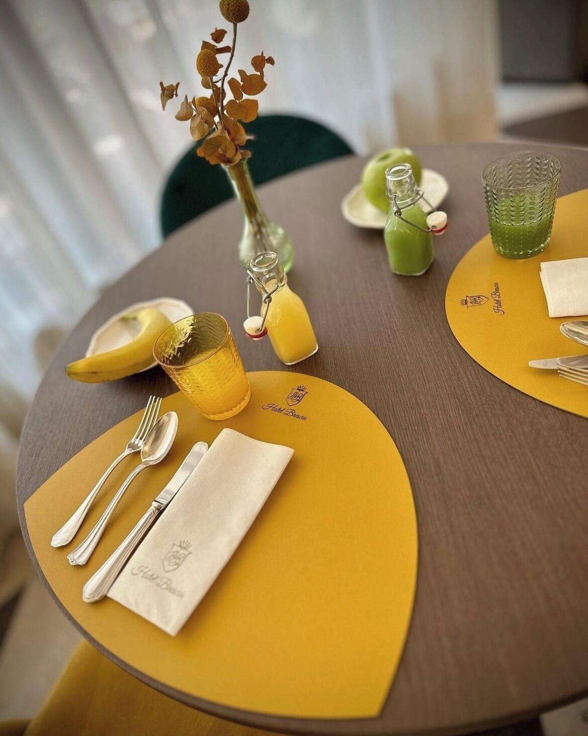 A round table with yellow placemats, a glass vase with dried flowers, a green drink in a glass, and a banana on the table.