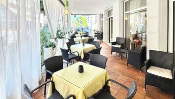 Een overdekt terras met tafels en stoelen, grote ramen en potplanten.