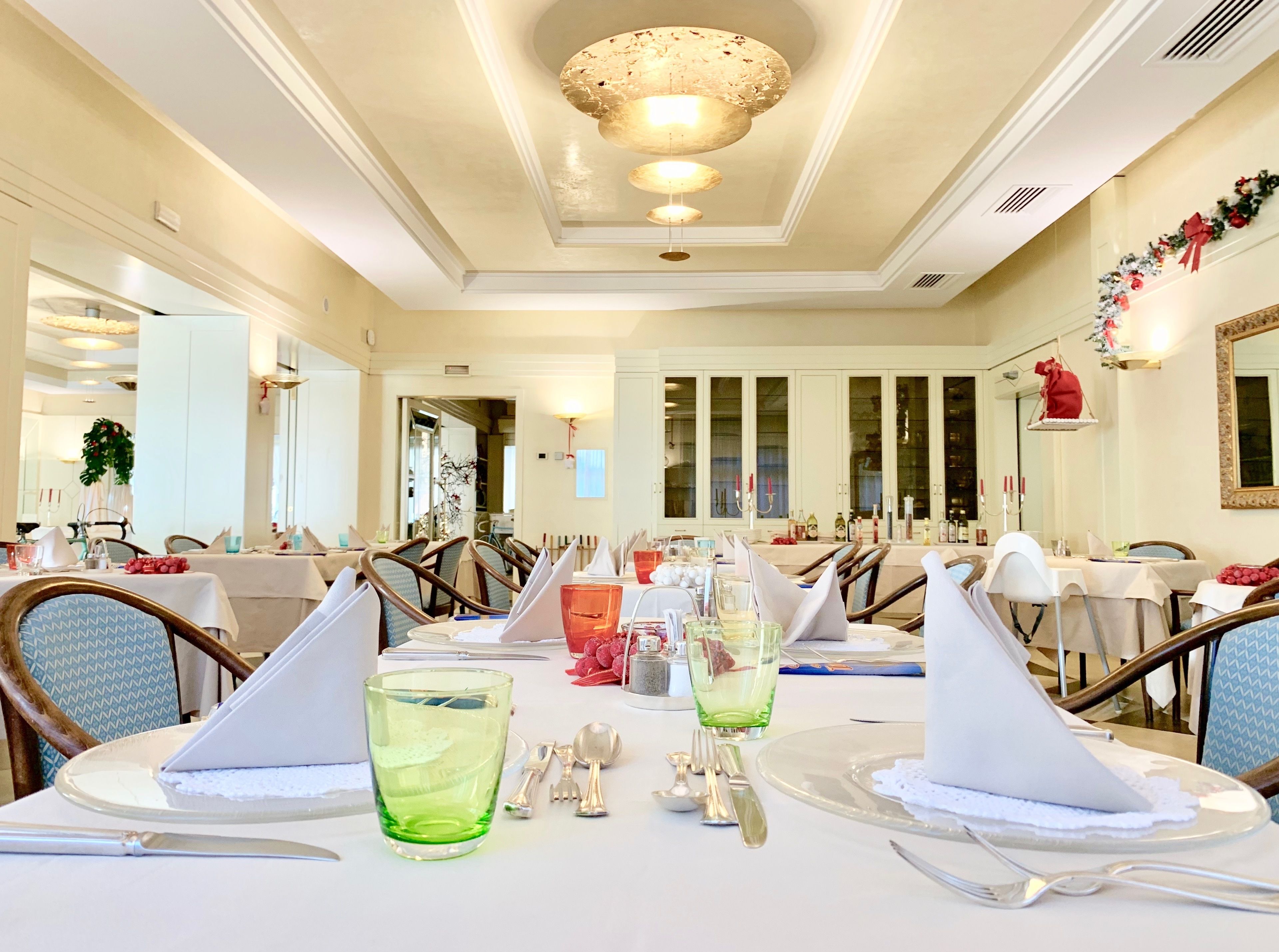 Une salle à manger bien éclairée, avec plusieurs tables dressées pour le service, ornées de nappes blanches, de couverts soigneusement disposés et de verres verts.