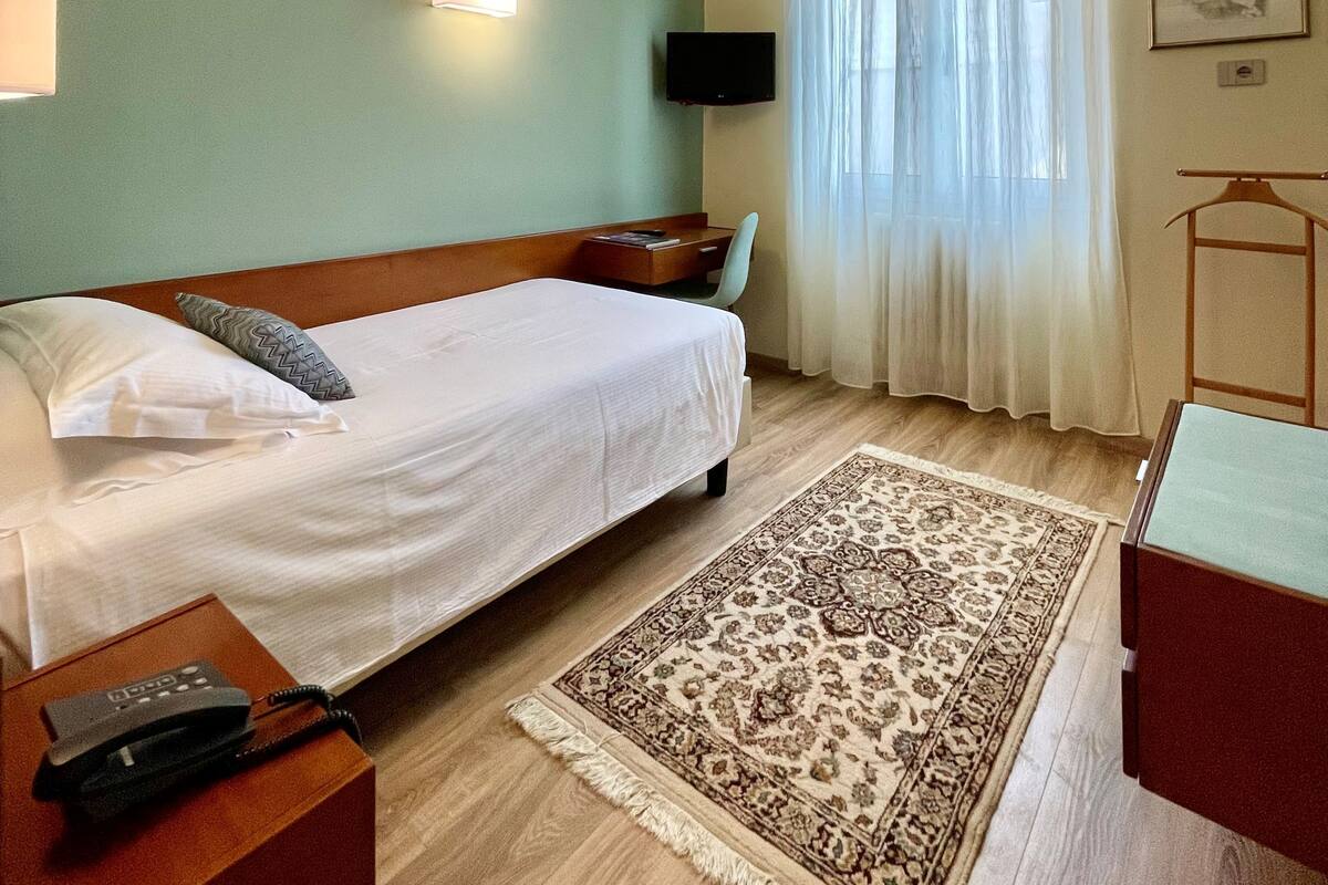 Hotelkamer met een bed, nachtkastje, bureau, stoel, televisie, raam met gordijnen en een kamerkleed met dessin.