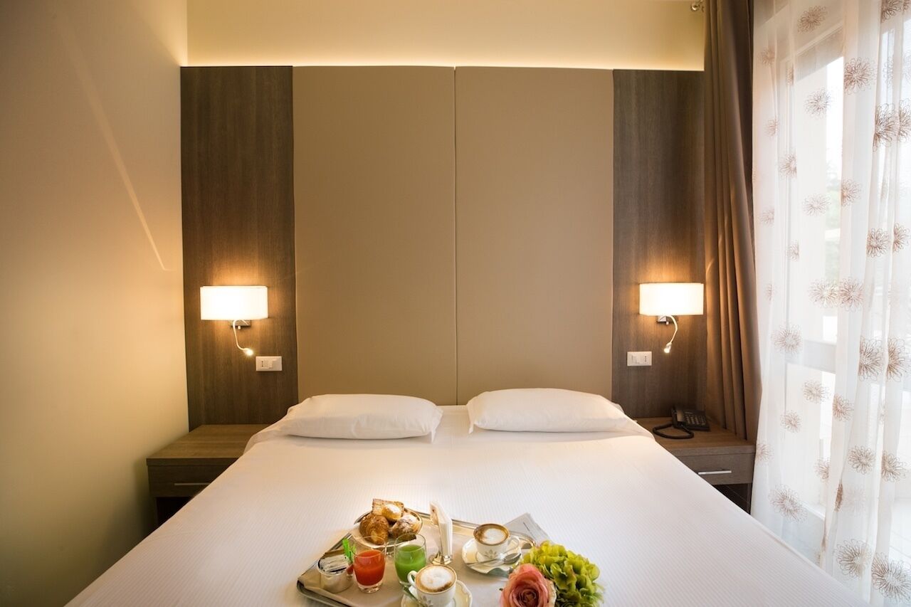Habitación de hotel con una cama grande, dos lámparas de noche, una bandeja con alimentos para el desayuno y un teléfono en la mesita de noche.