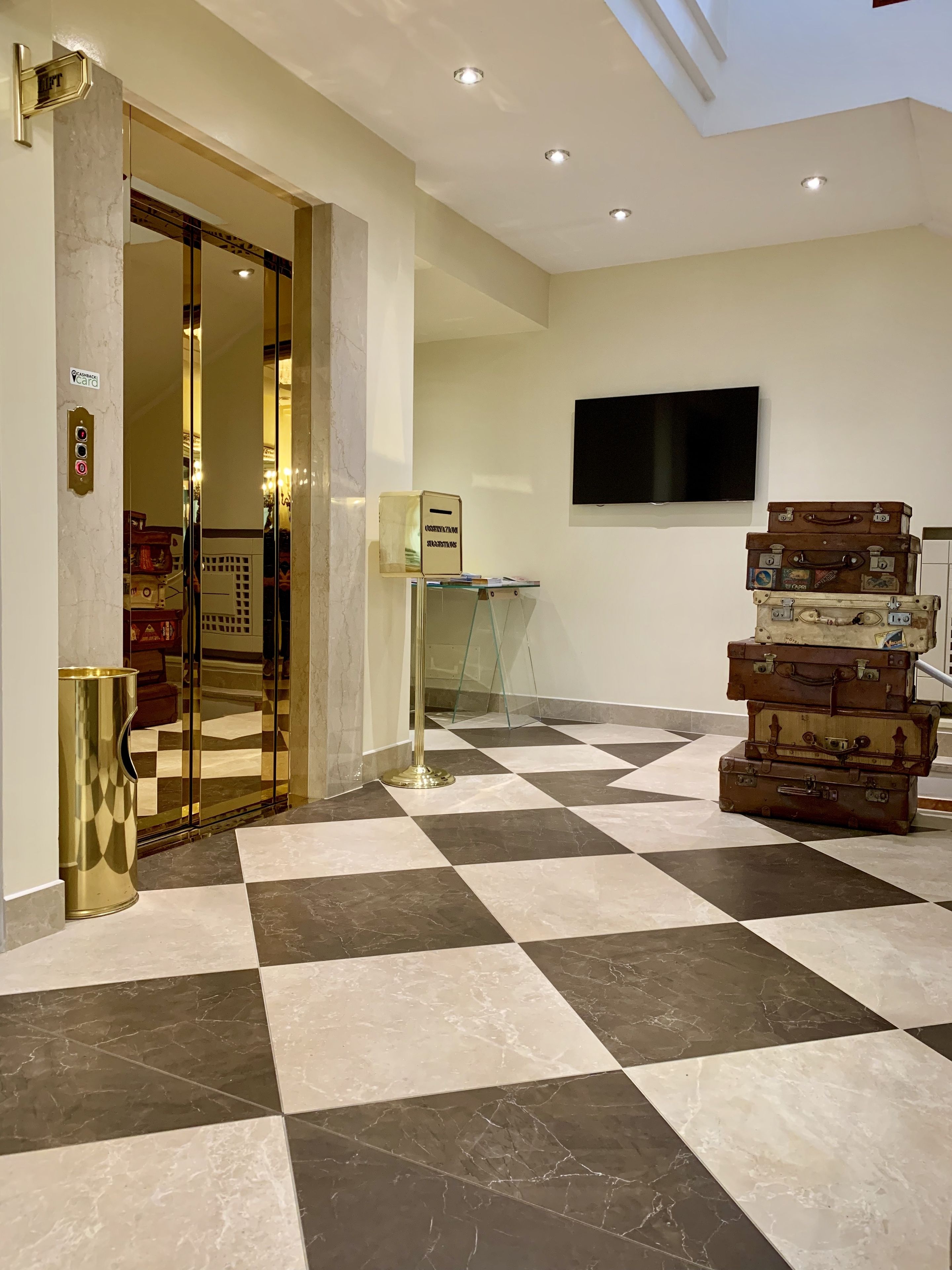 Un hall d’hôtel avec un sol à carreaux, un ascenseur en verre, un téléviseur fix é au mur et des valises empilées.