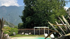 Una piscina con solárium de baldosas, rodeada de follaje y un pabellón blanco.