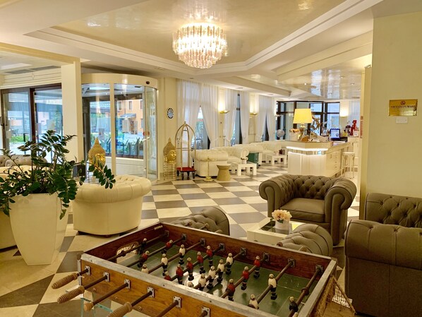 Un hall d’hôtel luxueux, avec un sol à carreaux, des fauteuils confortables, une table de baby-foot et un lustre.
