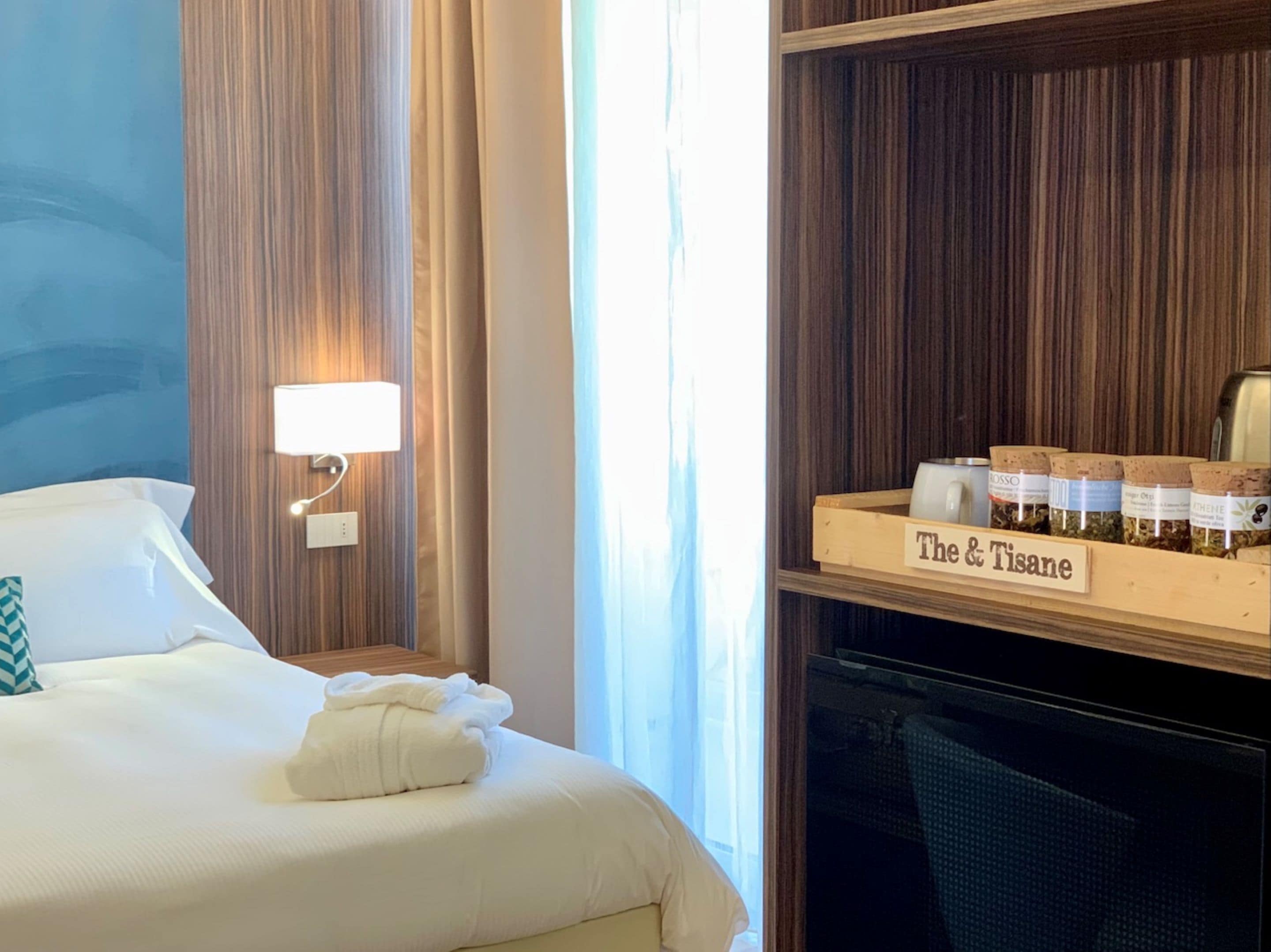 A hotel room with a neatly made bed, a bedside lamp, a shelf with jars labeled "The & Tisane", and a view of the outdoors through the window.