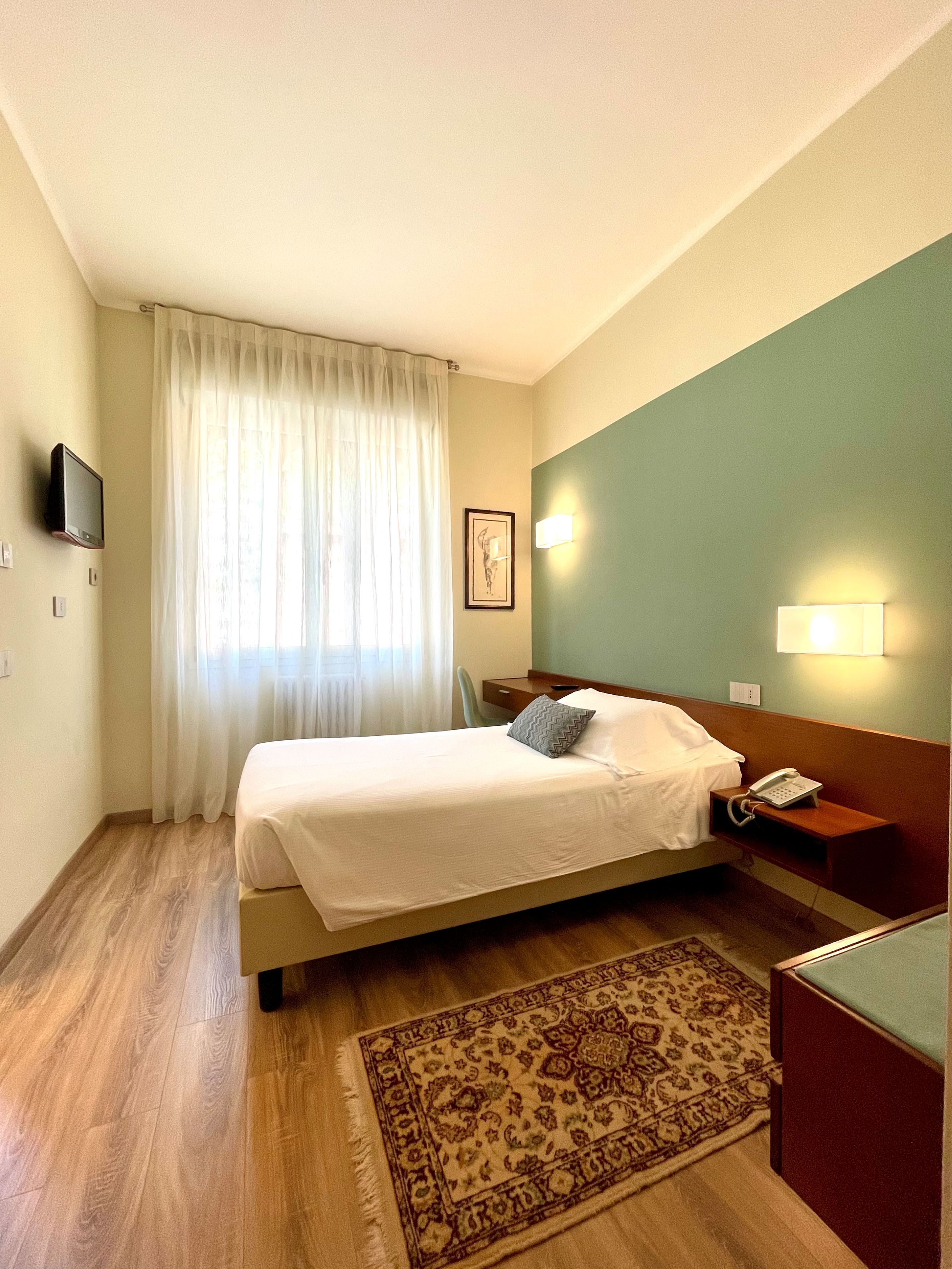 Une chambre d’hôtel avec un lit, un bureau, un téléphone, un tableau au mur et une fenêtre avec des rideaux.