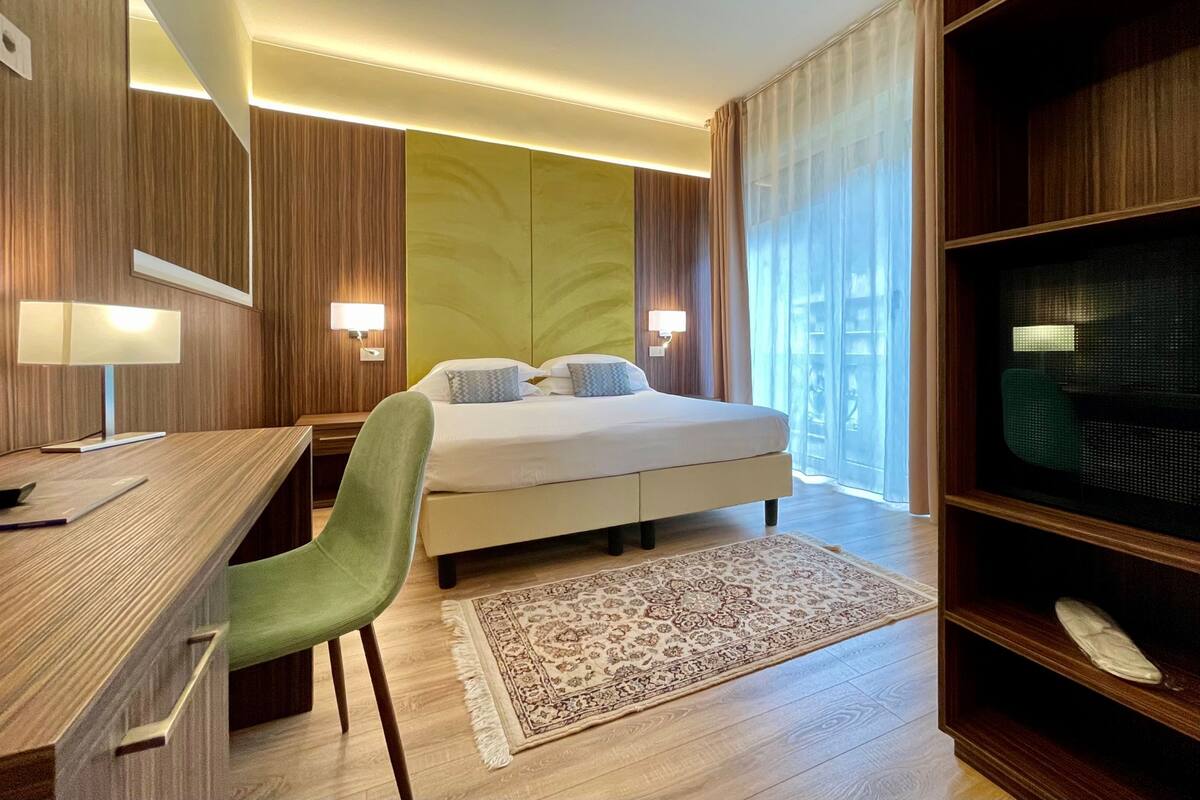 Een hotelkamer met een bed, bureau, stoel, televisie en een groot raam met gordijnen.