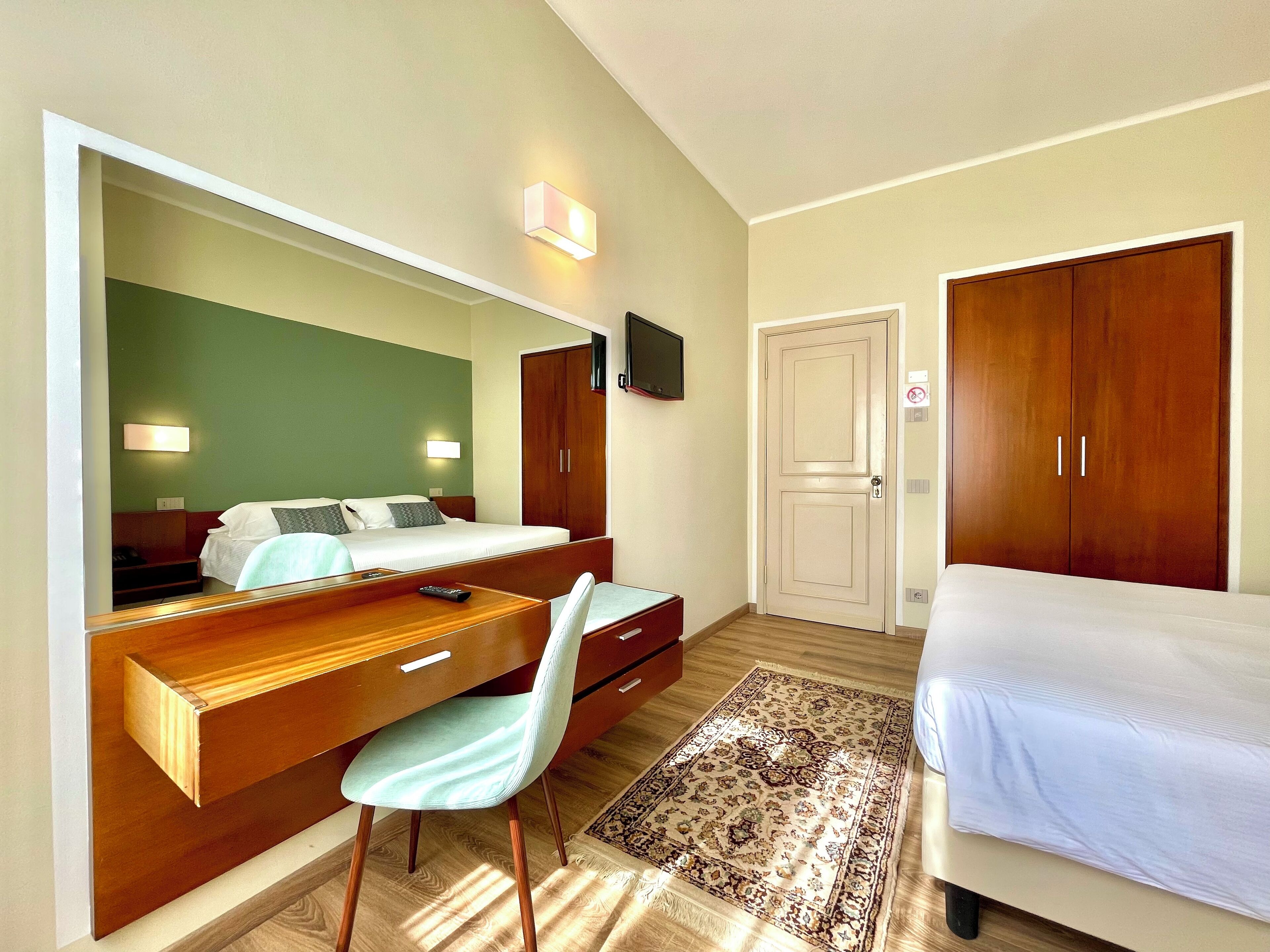 A hotel room with a bed, a desk with a chair, a large mirror, a wardrobe, and a television on the wall.