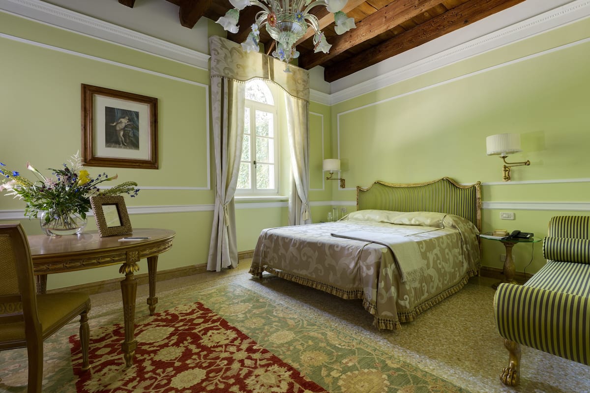 A room with a bed, a chair, a table with a vase and picture frame, a lamp, and a window with curtains.