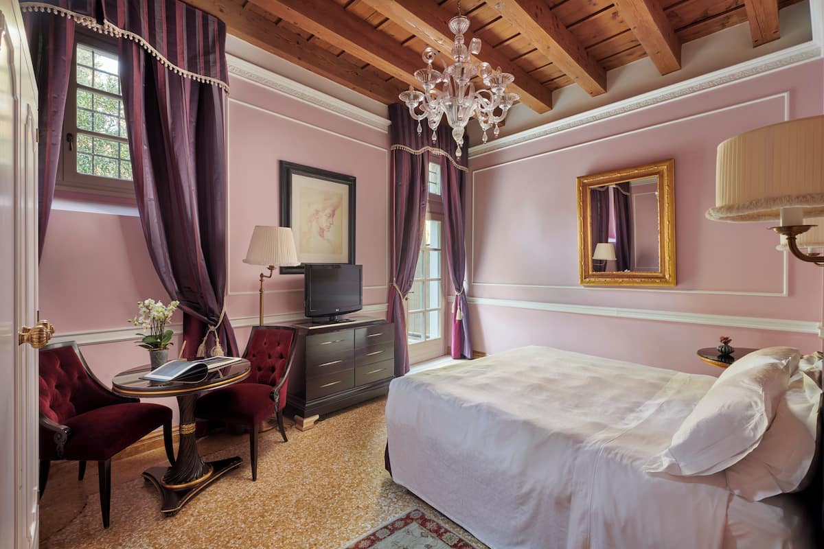 A bedroom with a bed, a TV, a desk, a chair, and a chandelier.