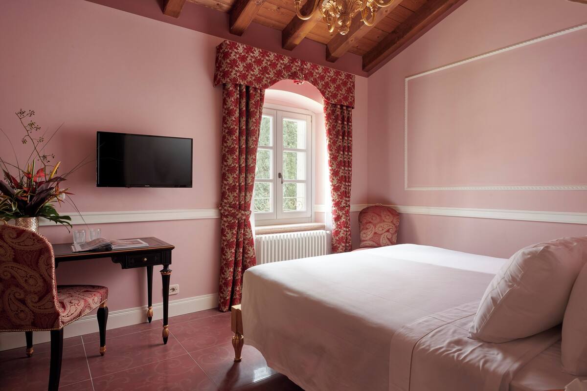 A bedroom with a bed, a desk with a chair, a television, and a window with floral curtains.