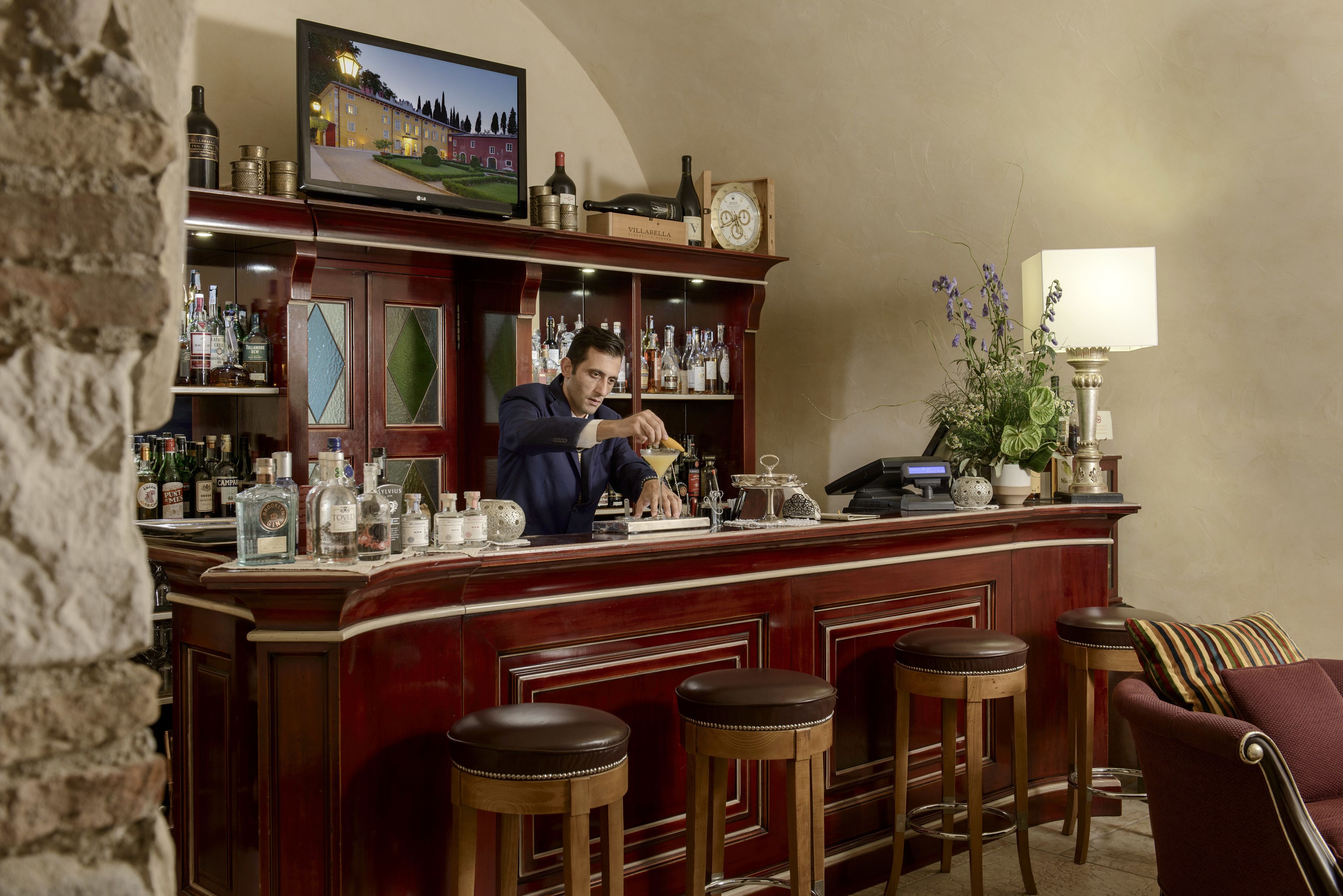 Zona de bar con un bartender, mostrador de madera, taburetes y un televisor que muestra un paisaje.
