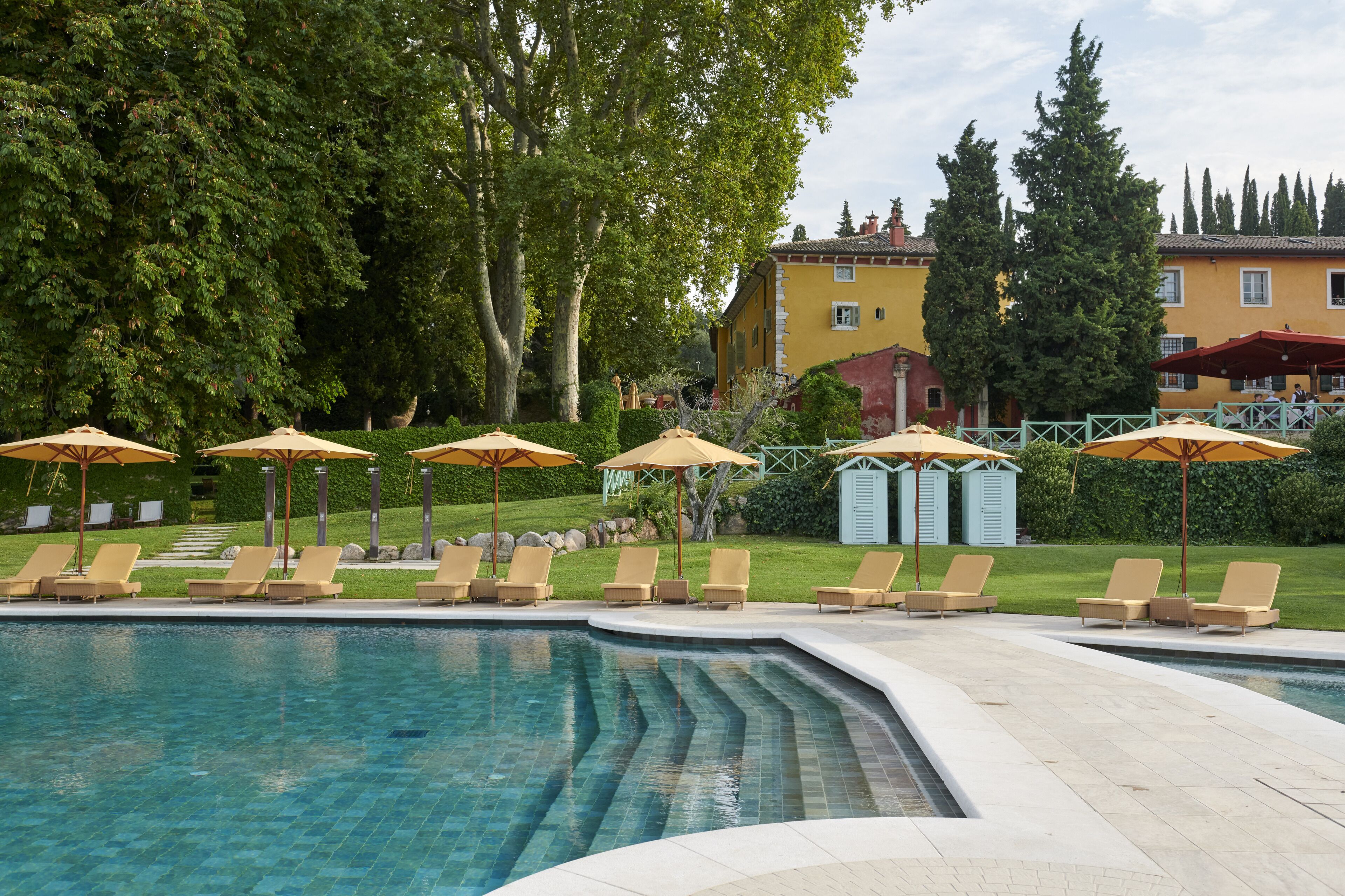 Ein Poolbereich mit Liegestühlen und Sonnenschirmen, umgeben von viel Grün und einem Gebäude im Hintergrund.