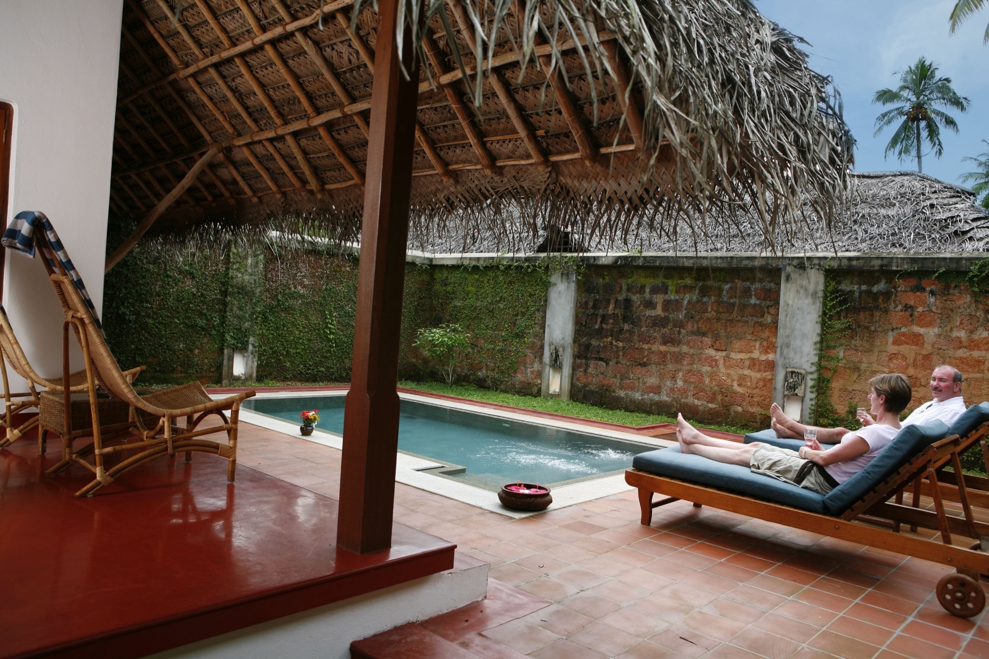 Zona junto a la piscina con sillas de descanso, techo de paja y pared de ladrillo con plantas.