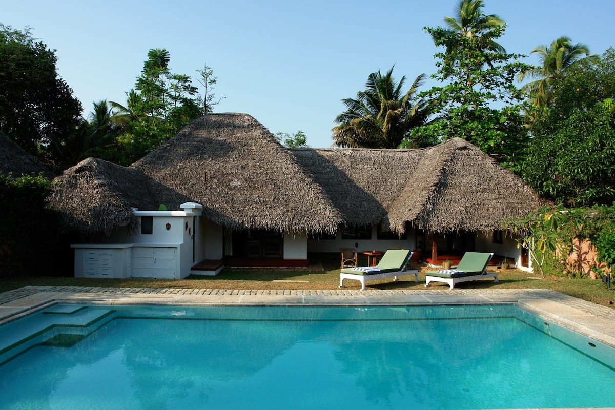 A pool with lounge chairs, a thatched-roof building, and lush greenery.